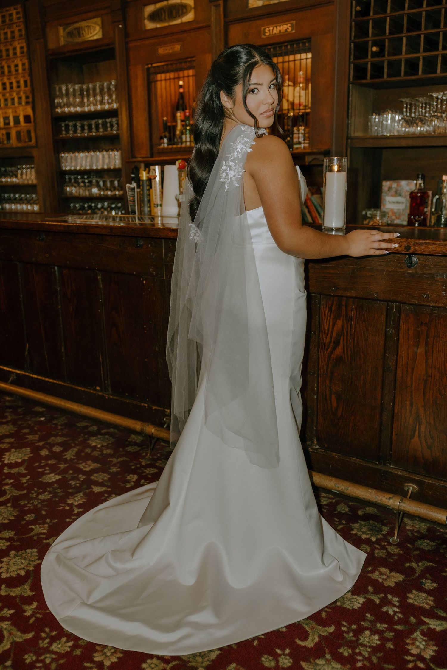 A flowing white wedding dress train extends across a vintage bar's carpeted floor.