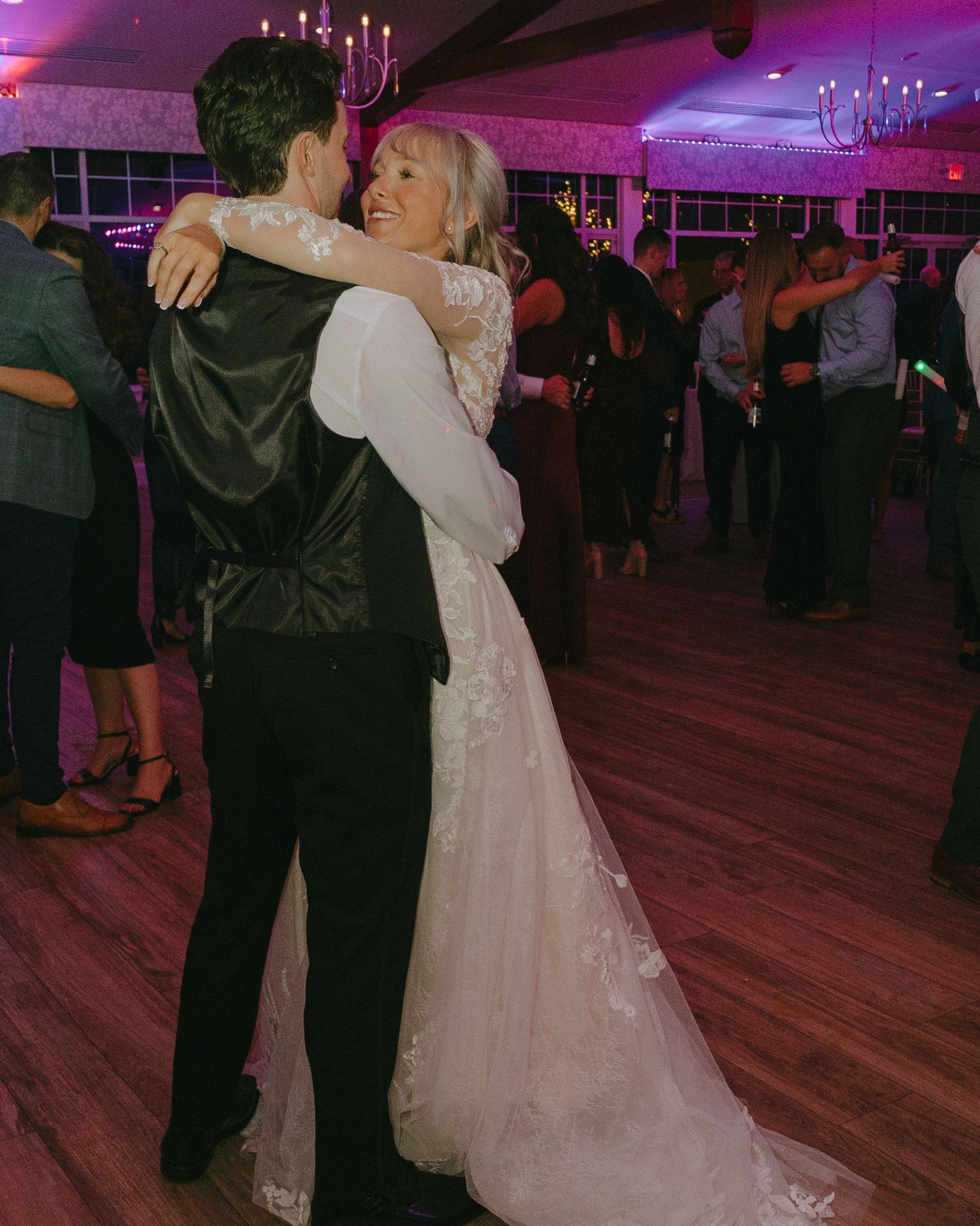 Newlyweds share their first dance on wooden floor under purple lighting at evening reception.