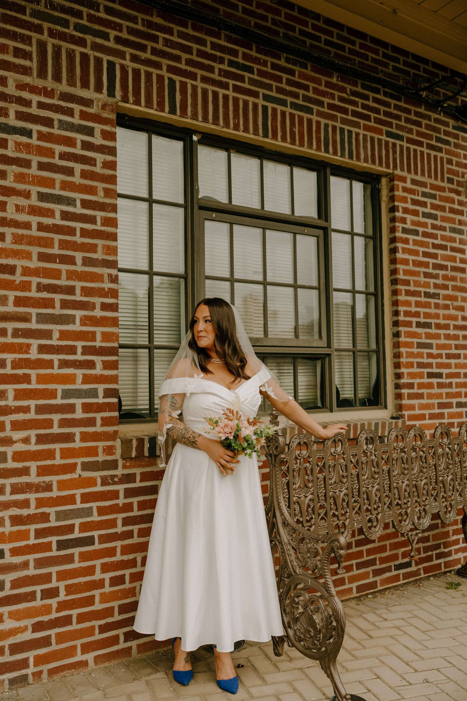 A short white wedding dress is showcased against an ornate iron bench and brick wall backdrop.