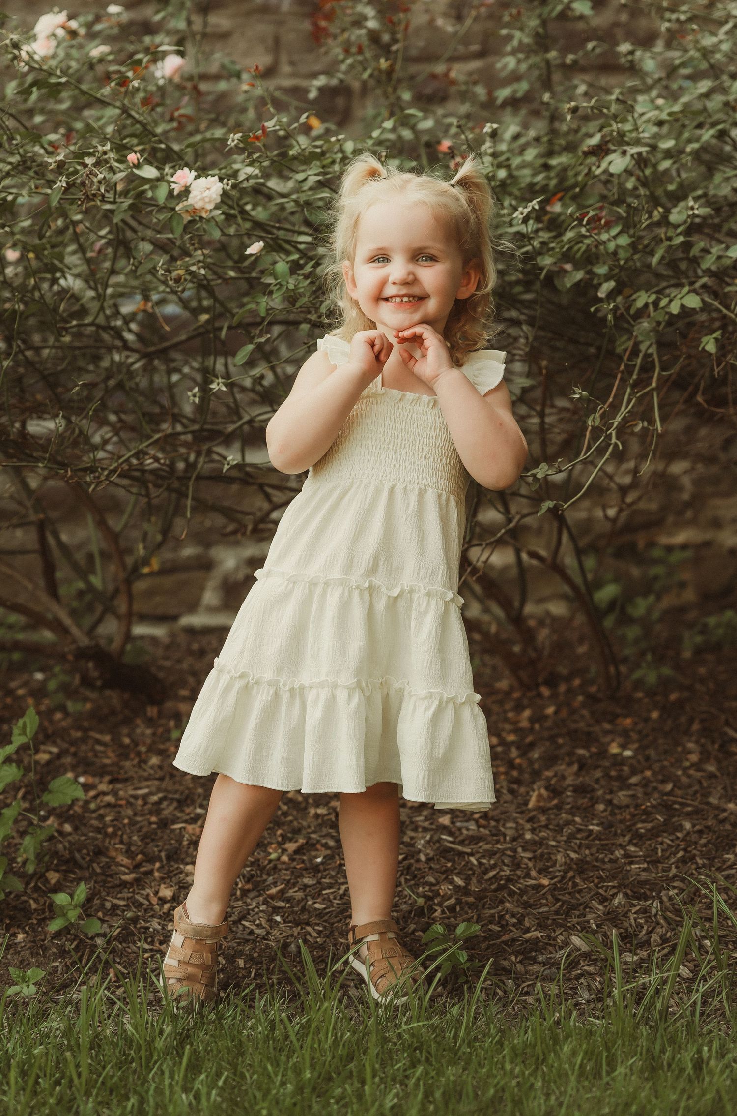 Person in cream colored dress poses cheerfully against garden background with stone wall.