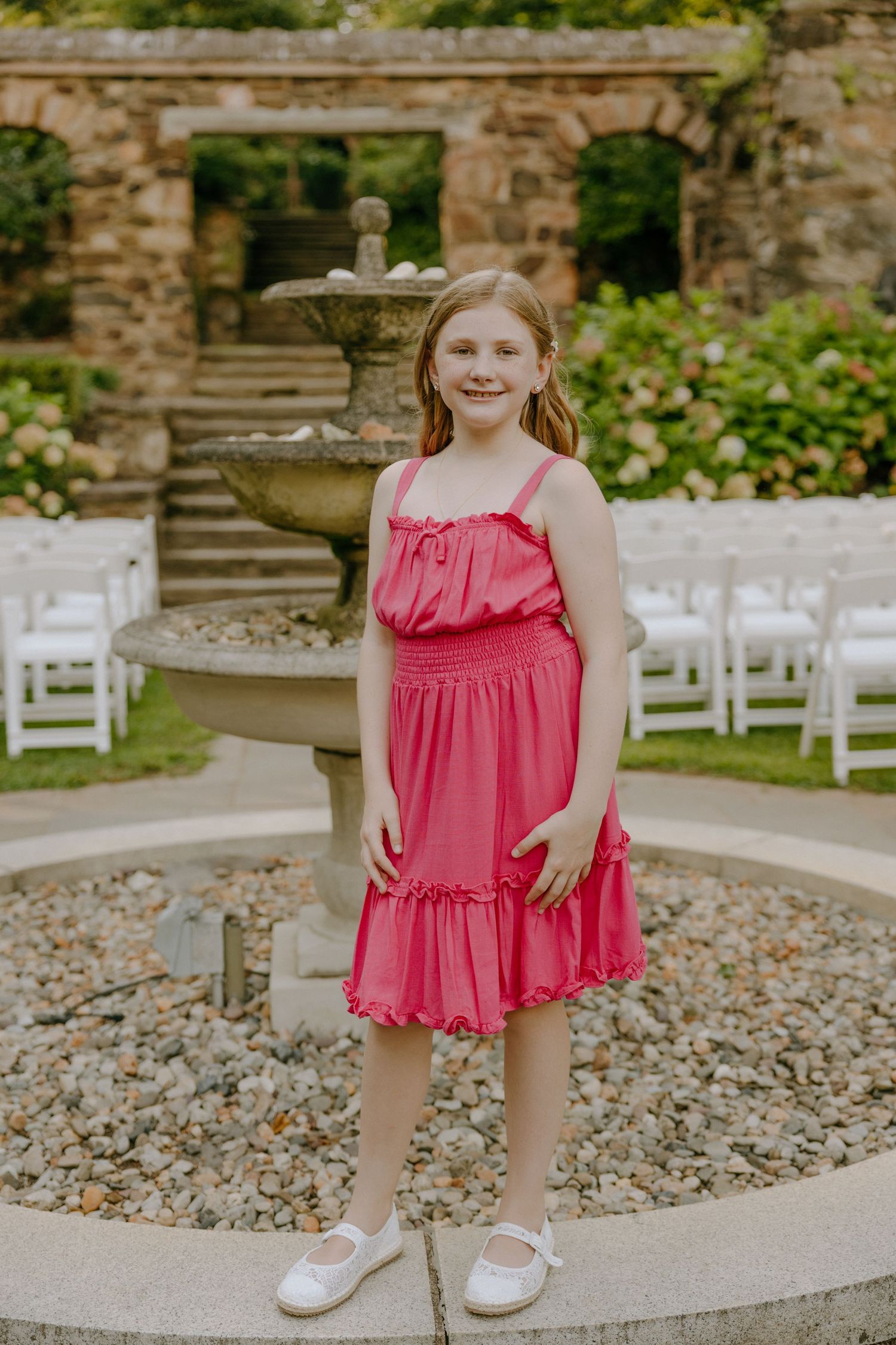 Person in coral ruffled dress poses on circular stone platform near white garden chairs.