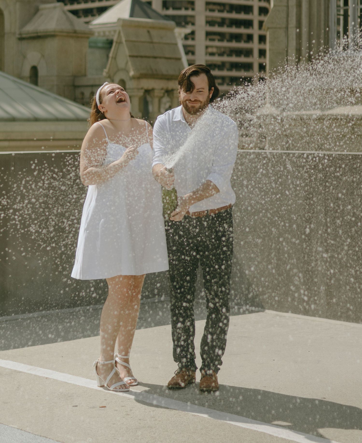 A couple in white outfits plays in water sprinklers on a sunny day outside a building.