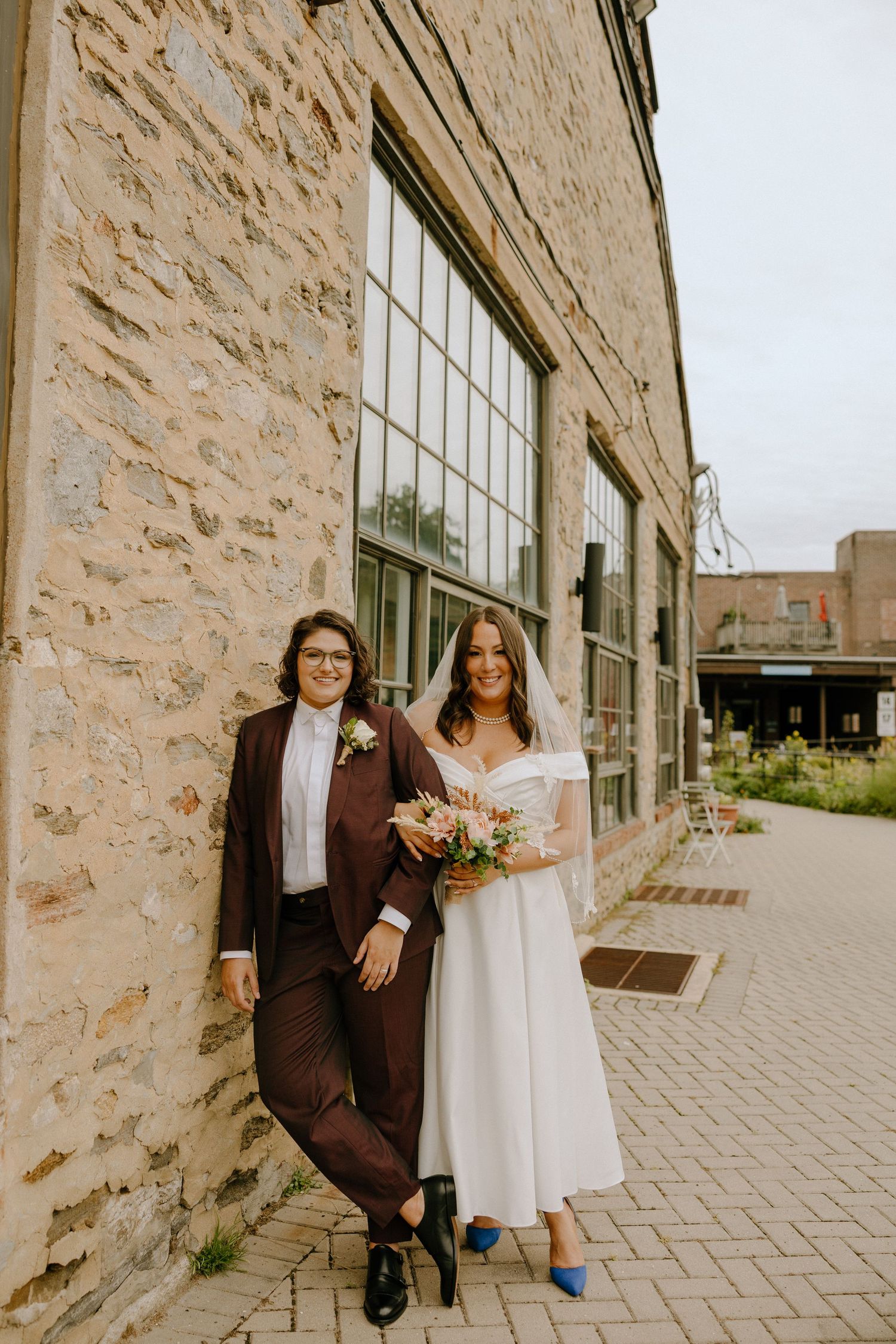 Wedding couple poses along a historic stone building with large windows and paved walkway.