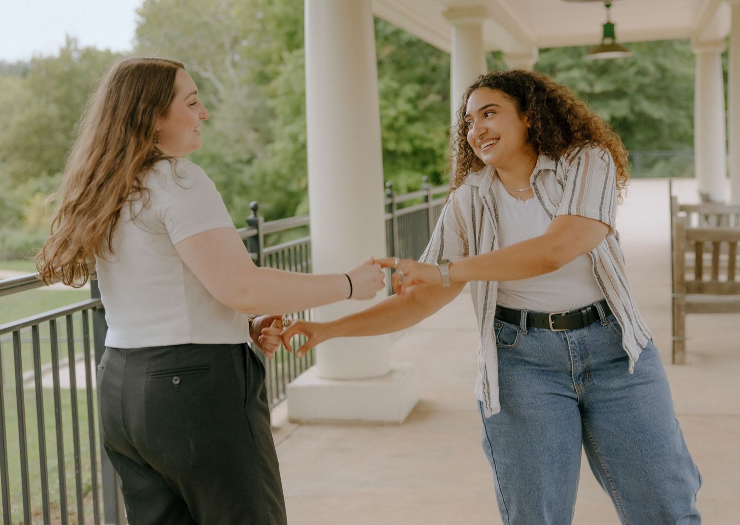 Two friends holding hands and laughing on a covered porch with columns.