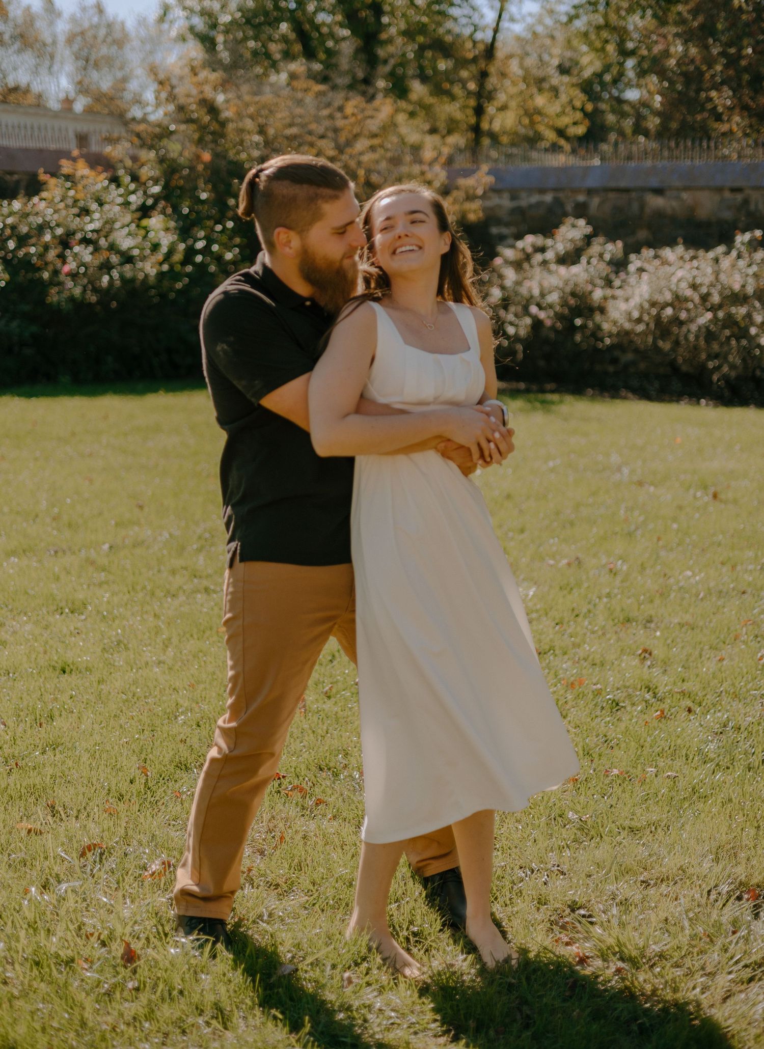 A couple embraces in a sunny garden with golden sunlight and blooming flowers in the background.