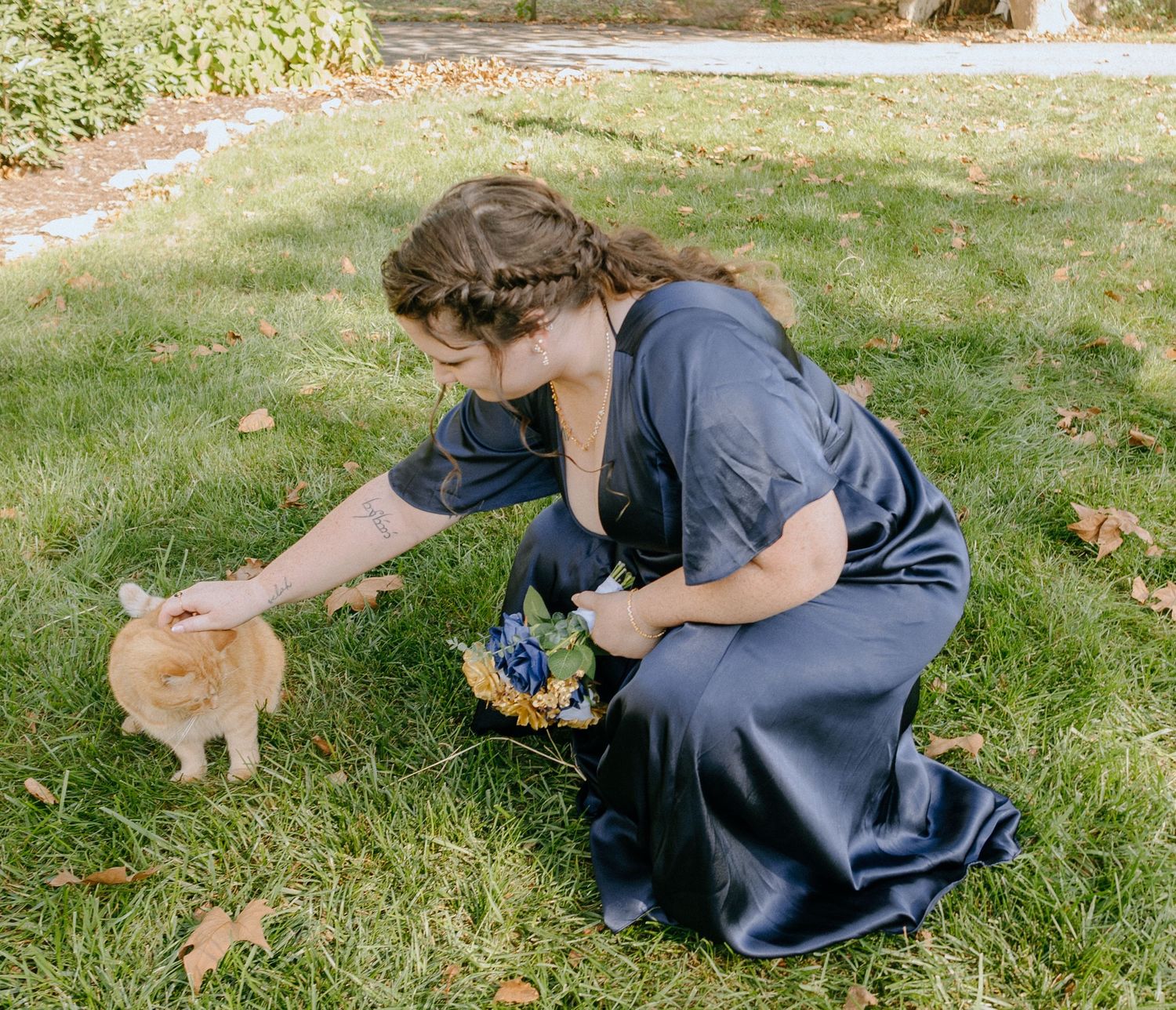 A person in a navy blue dress sits on grass petting an orange cat while holding flowers.