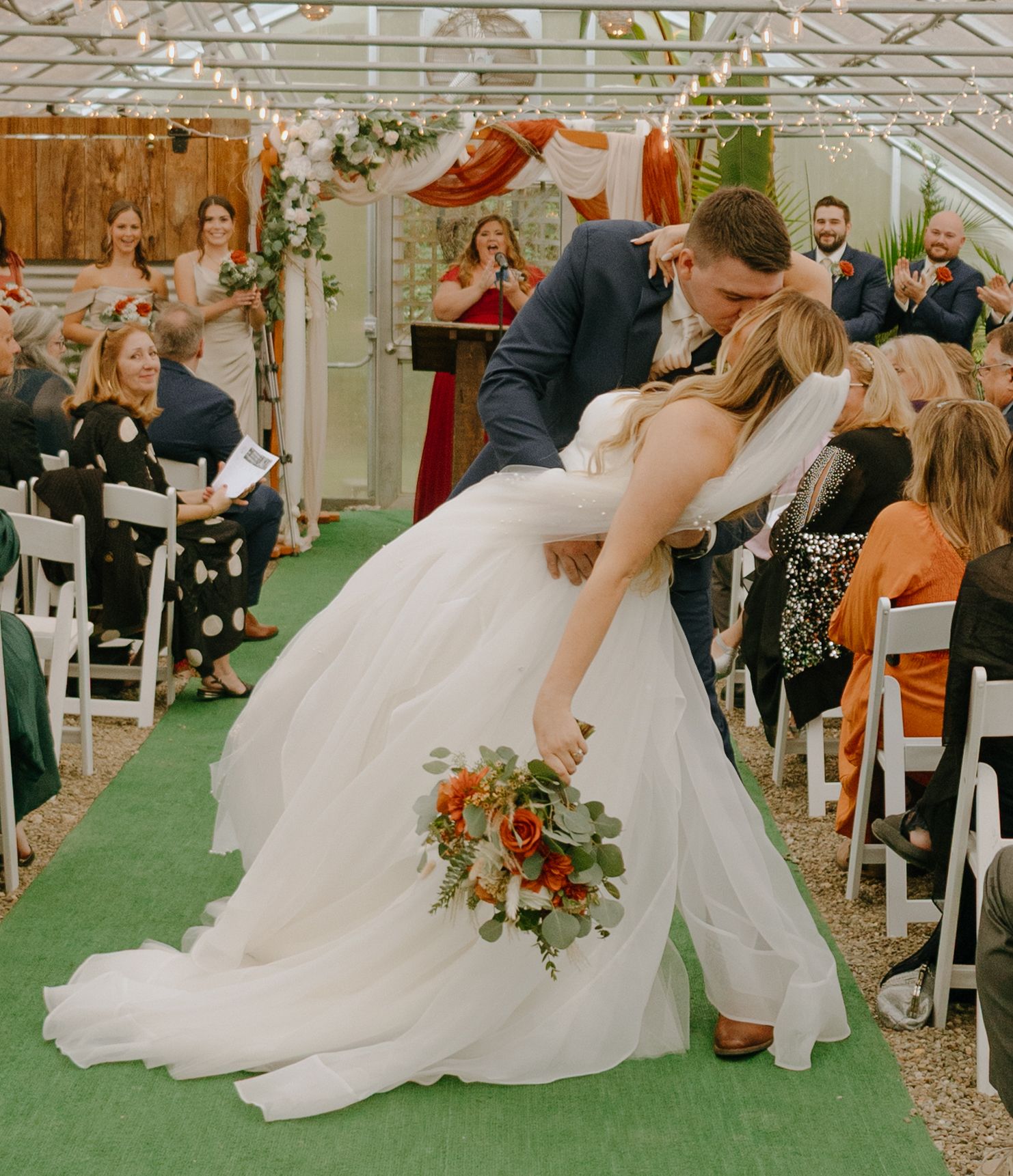 Bride and groom share a romantic dip during their outdoor wedding ceremony on artificial turf with guests watching.