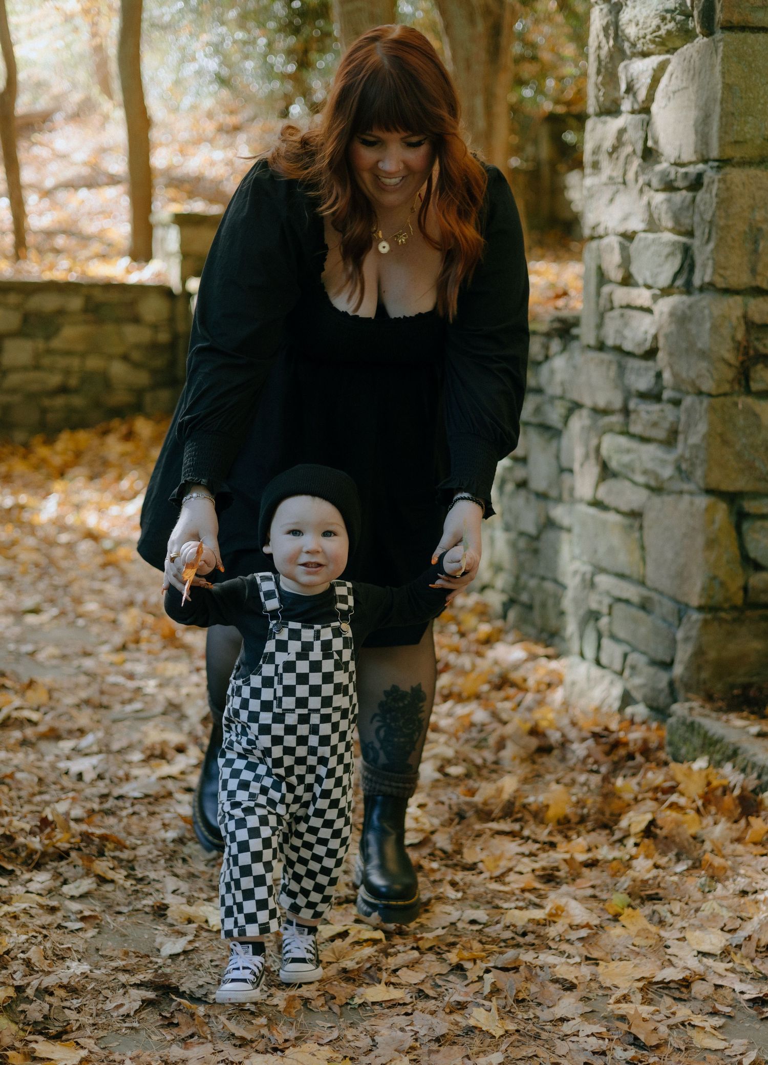 A toddler in checkered overalls takes first steps on autumn leaves while being supported on a stone path.