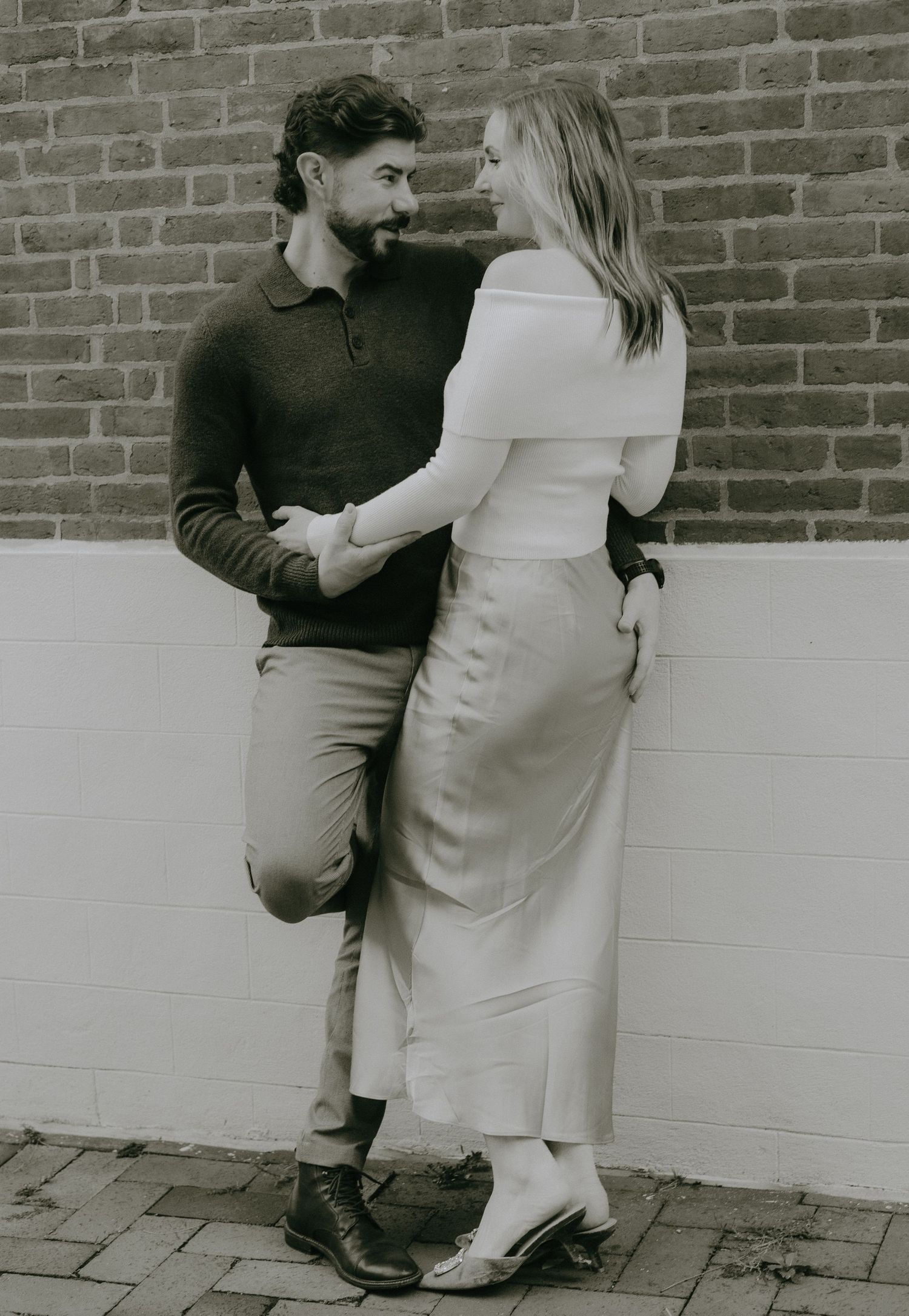 A romantic black and white photo of a couple in elegant attire standing close together against a brick wall.