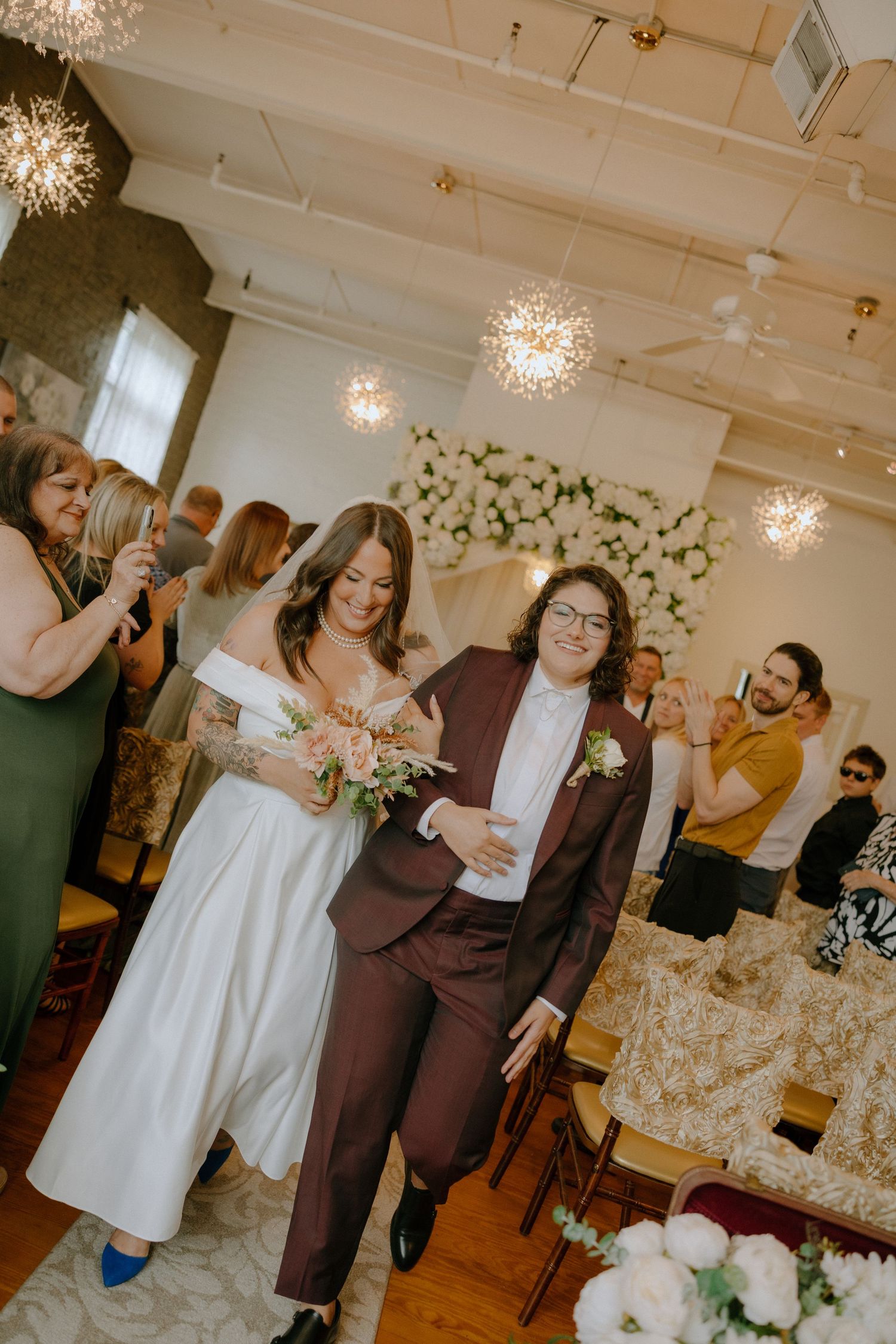 Wedding celebration in a bright venue with chandeliers as guests socialize near floral decorations.