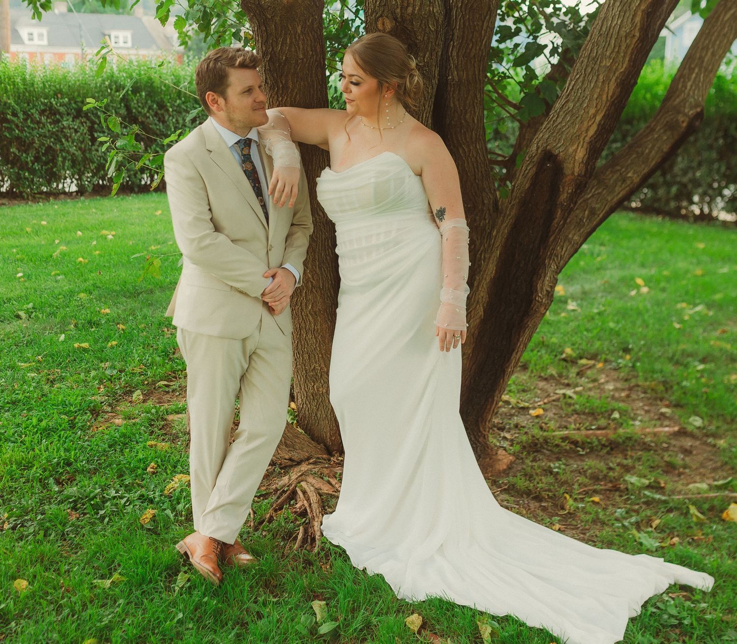 A couple in wedding attire sharing romantic moments under lush green trees in an outdoor garden setting.