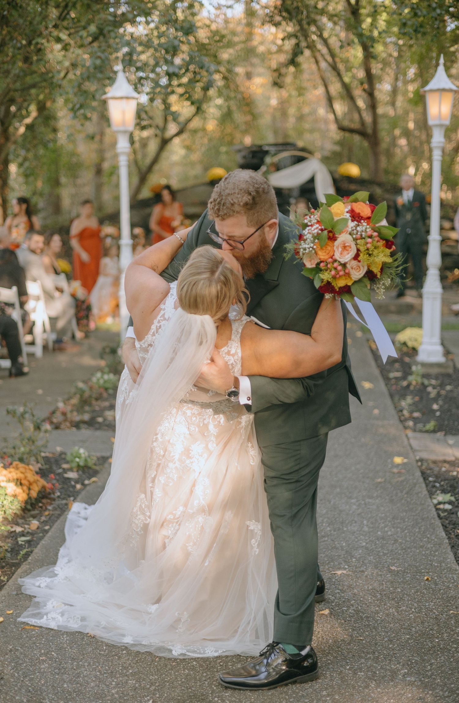 A couple shares a romantic moment on their wedding day along a tree-lined path with decorative lamp posts.
