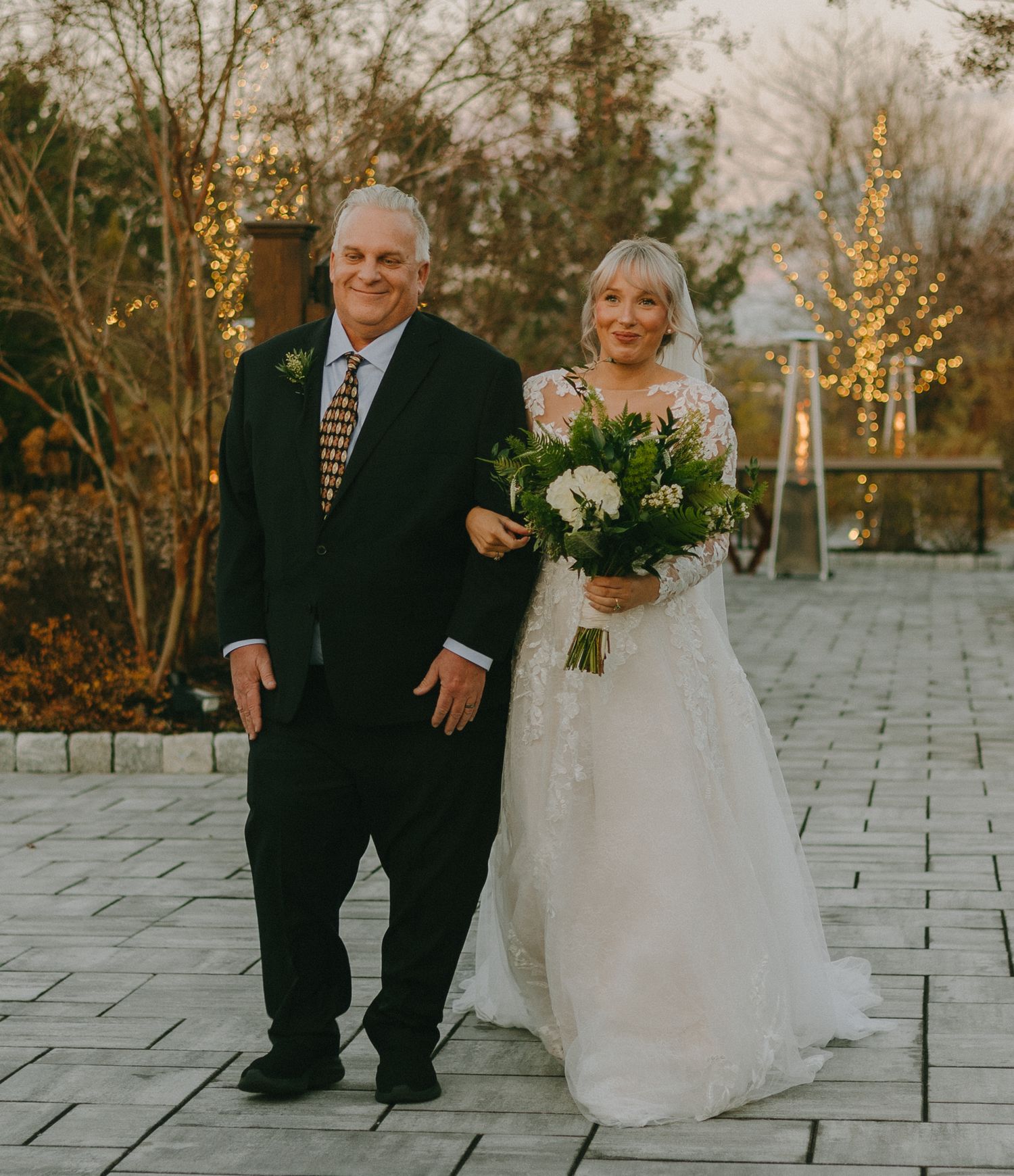 A couple in wedding attire poses together on a stone patio with autumn foliage in the background.