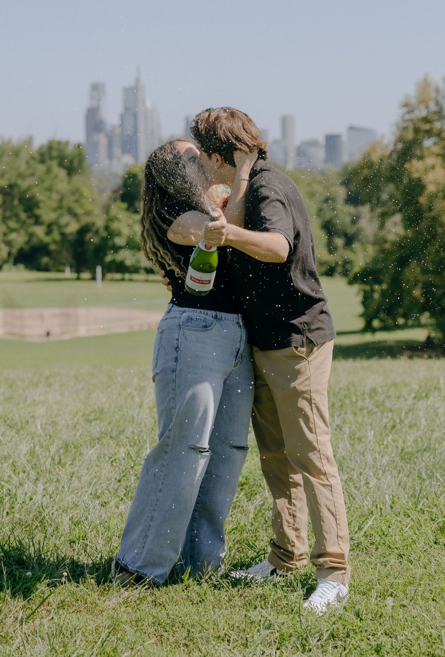 Celebratory moment with champagne spray in a sunny park with city buildings in the distance.