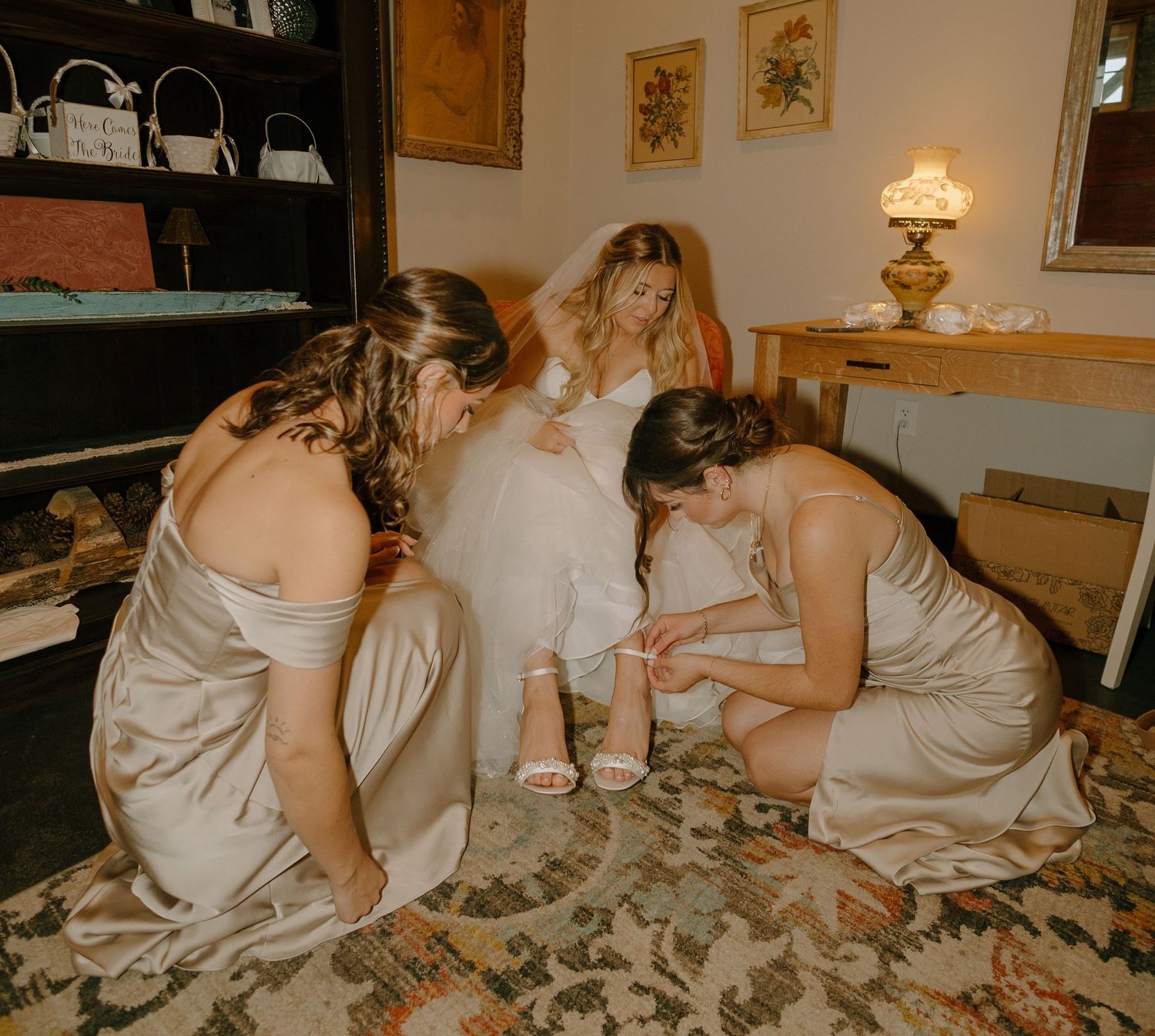 Three bridesmaids in champagne dresses help the bride with her gown while sitting on a patterned rug.