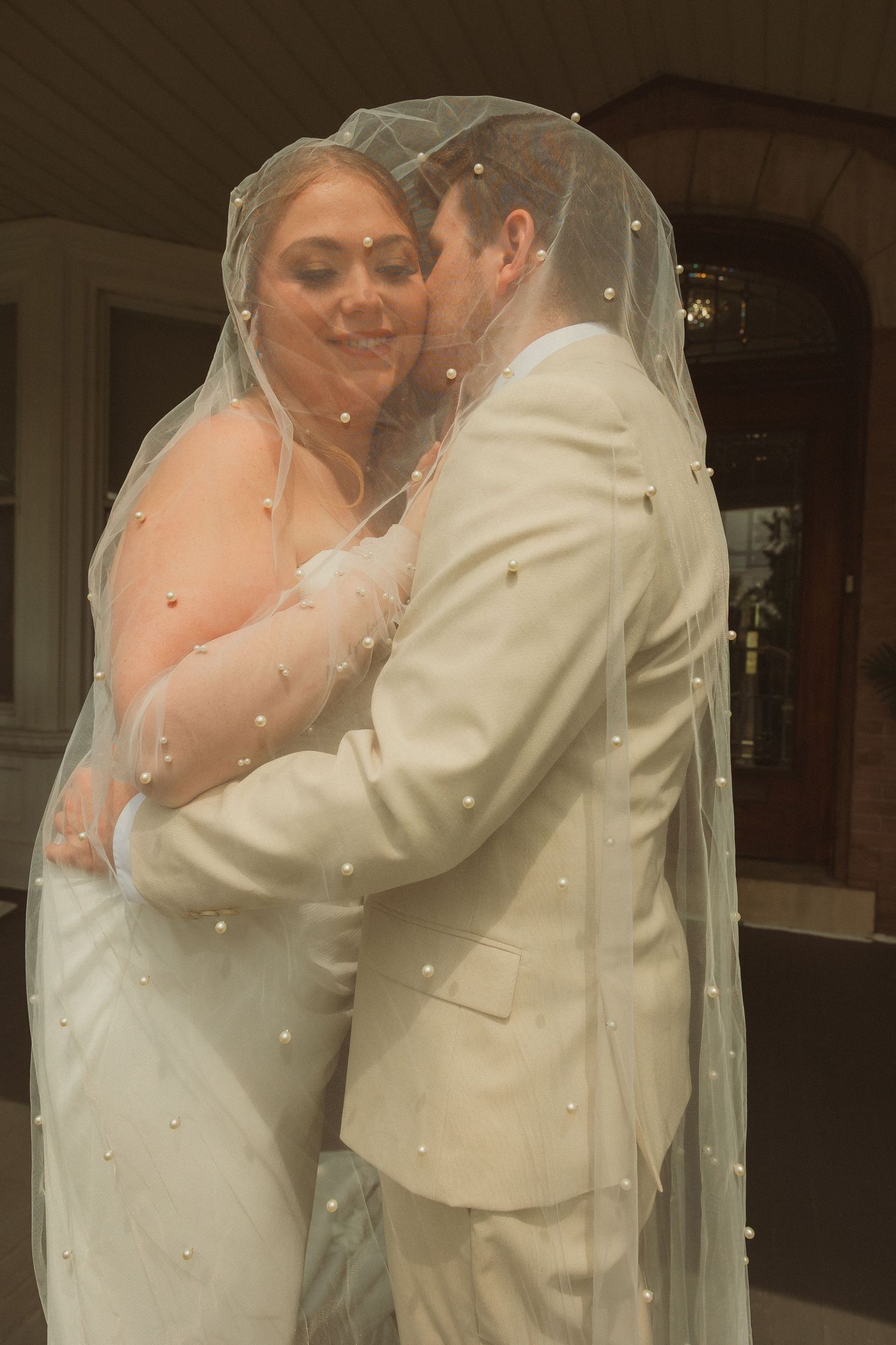 Romantic wedding portrait of couple embracing under dotted veil in formal attire.