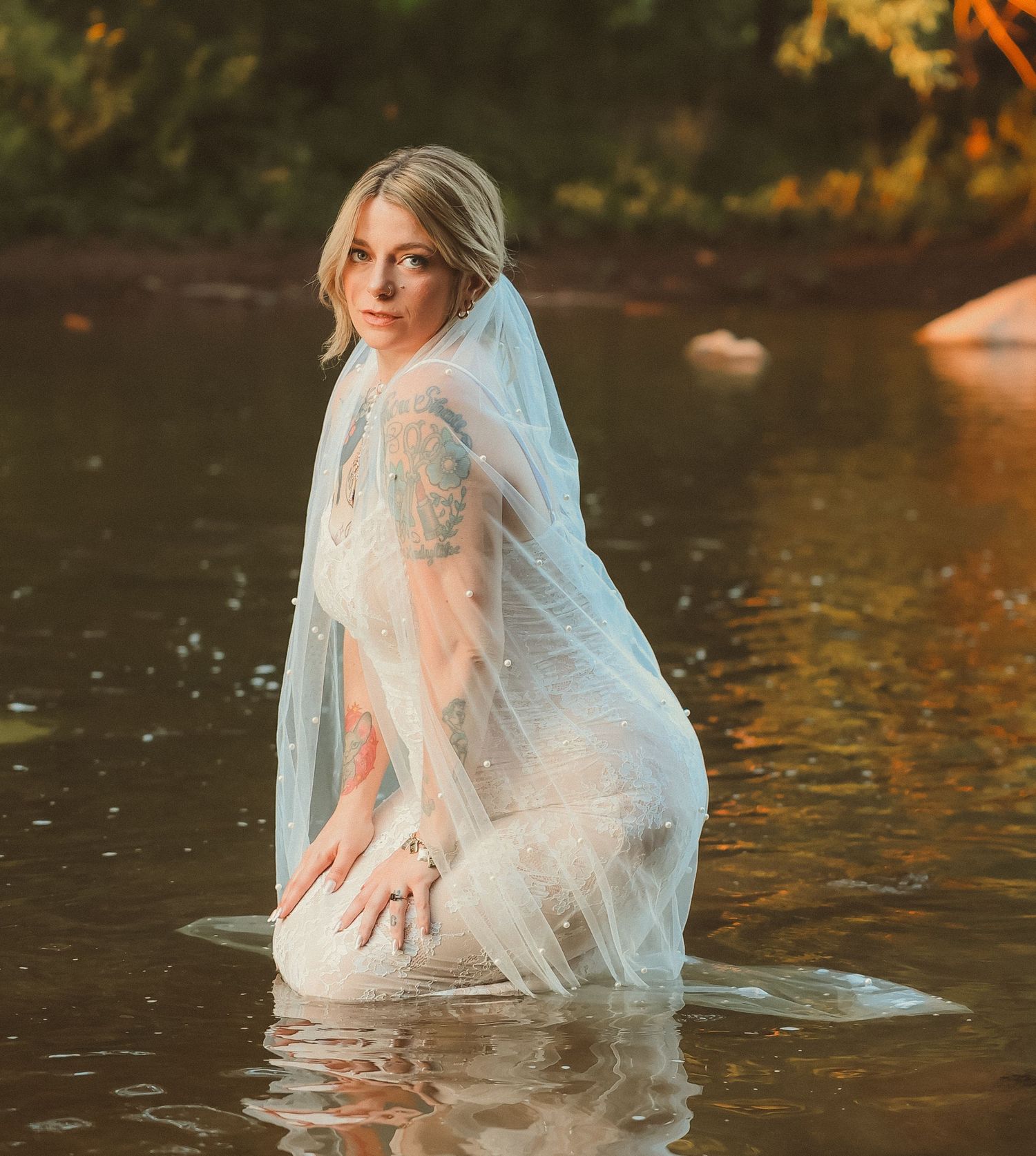A person in a white lace wedding dress and veil poses gracefully in river water.
