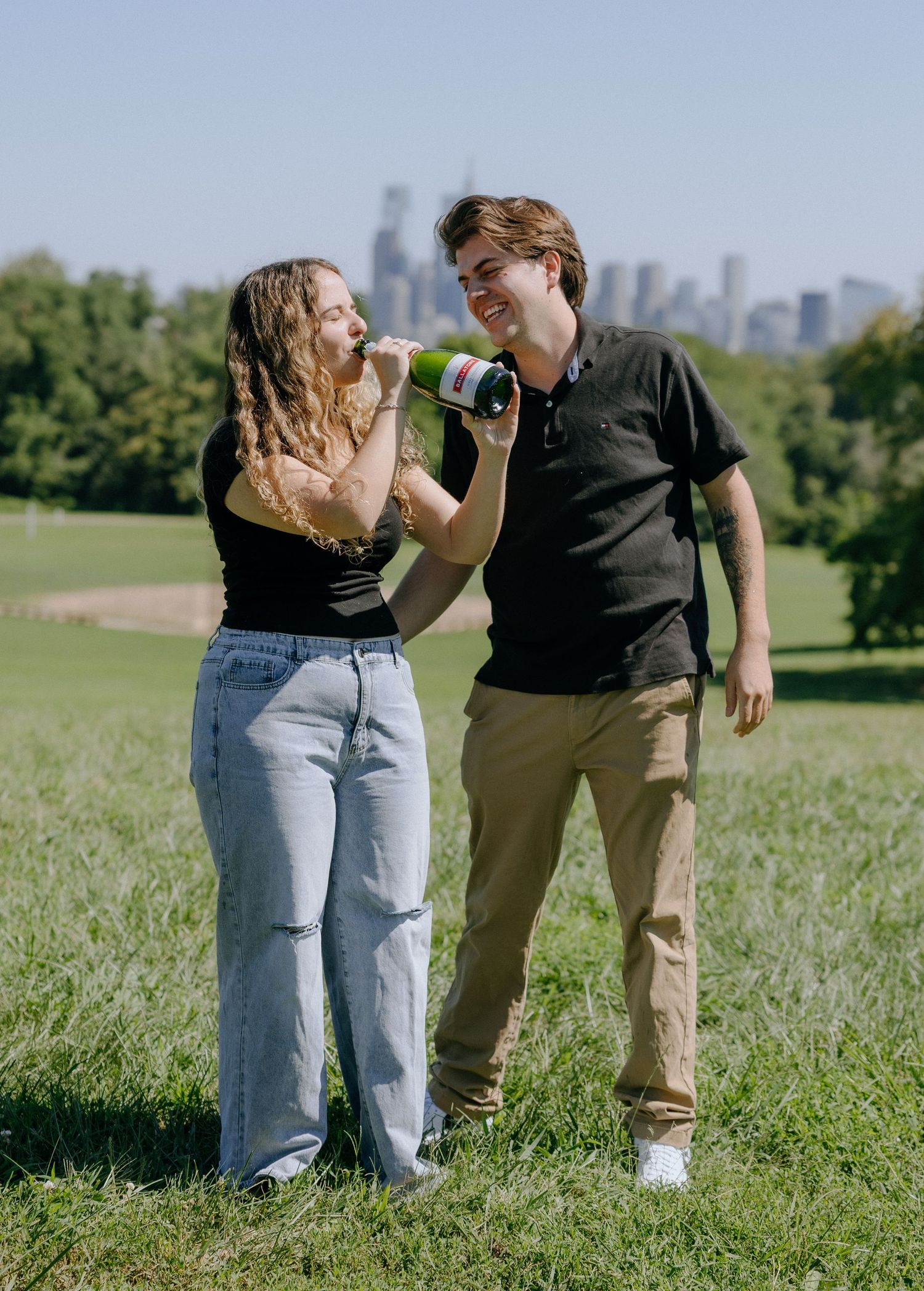 Candid moment of two people laughing and celebrating with champagne in a grassy park with city skyline visible.