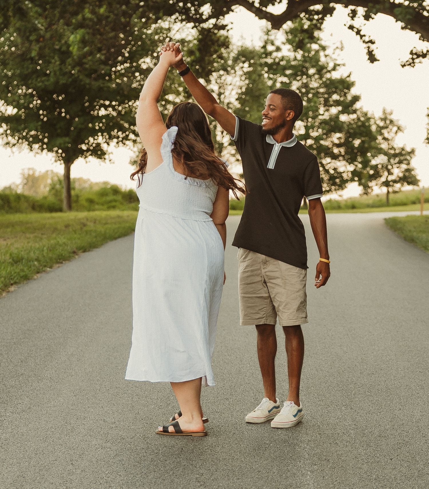 A couple in a white dress and khaki shorts pose playfully together on a country road at sunset.