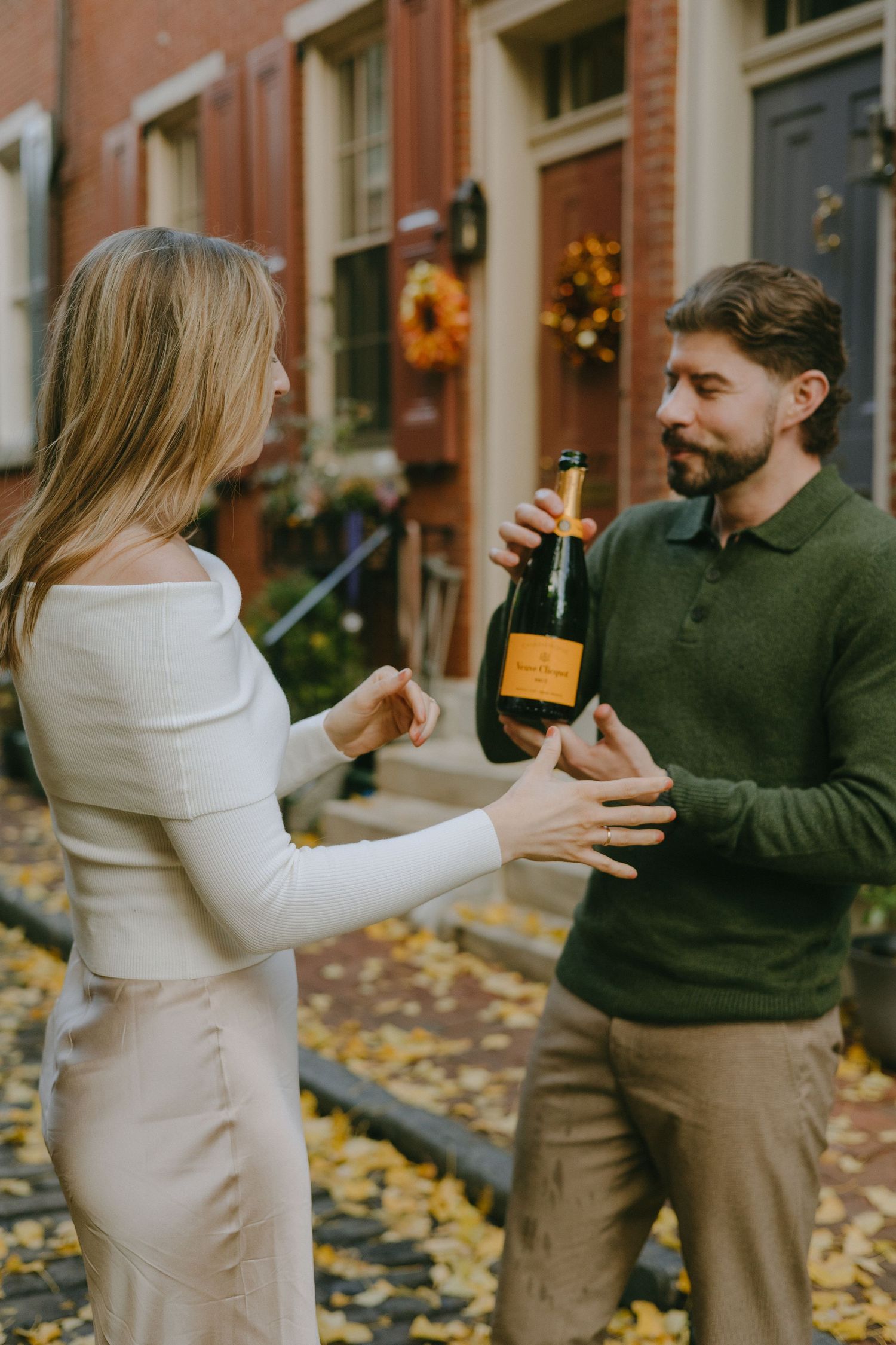 A couple celebrates with champagne on a fall day outside a brick building with autumn leaves on the ground.