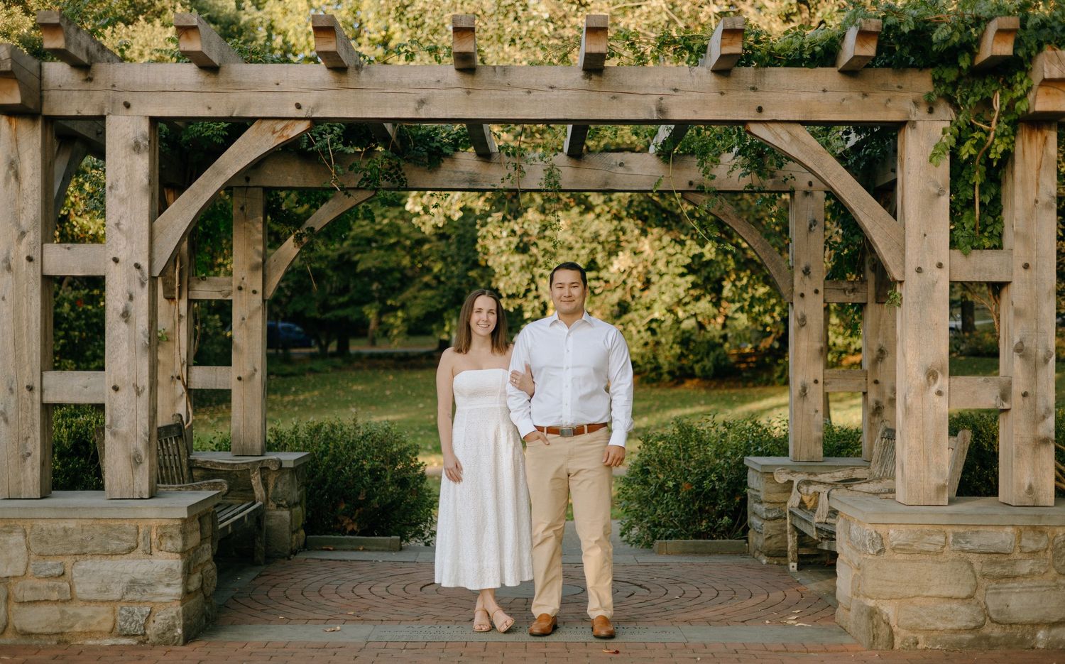 A rustic wooden pergola walkway with stone pillars in a garden setting during golden hour lighting.