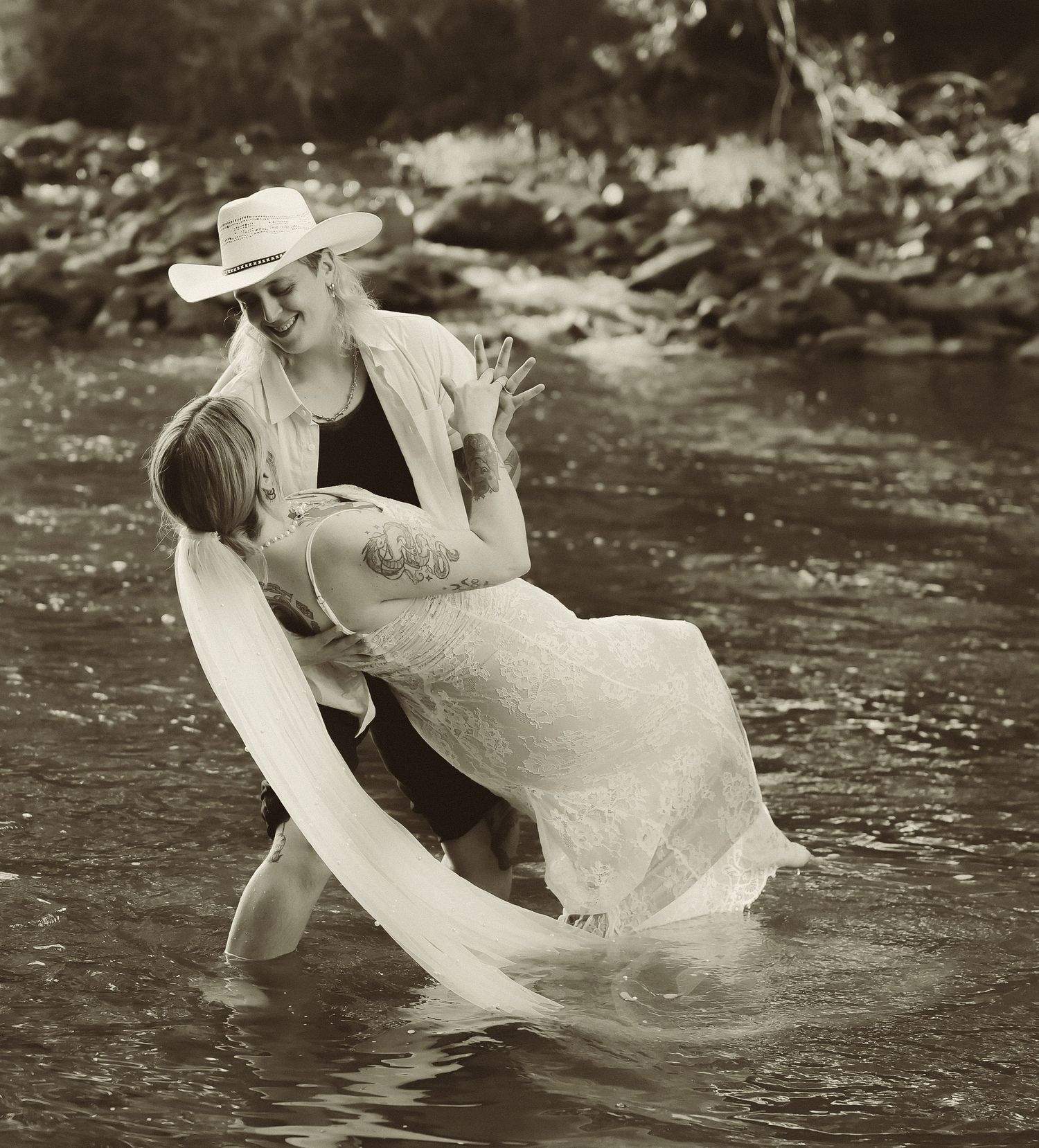 A black and white photo captures a playful moment in a flowing river during a wedding shoot.