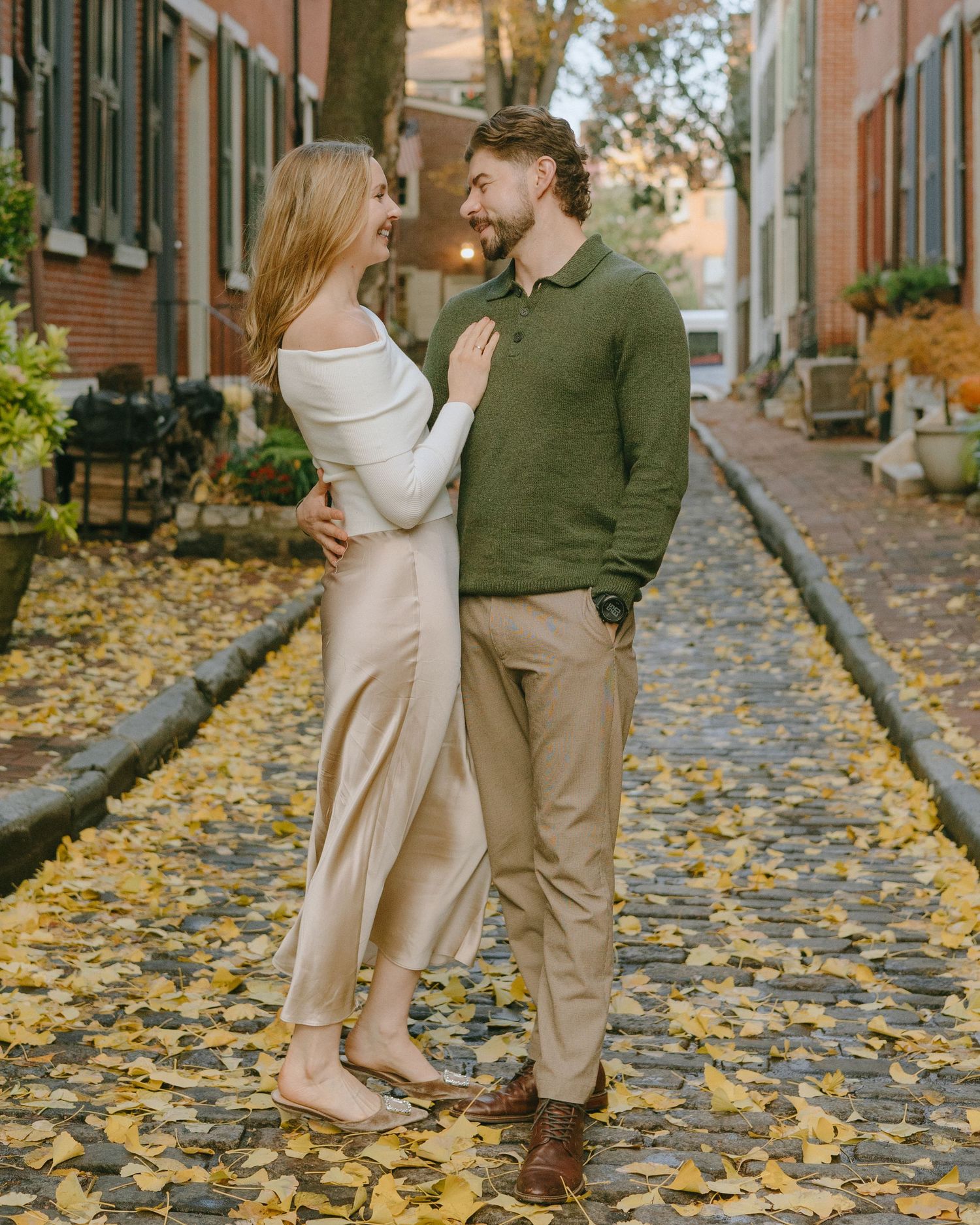 A couple embraces on a cobblestone street covered in golden fall leaves between brick buildings.