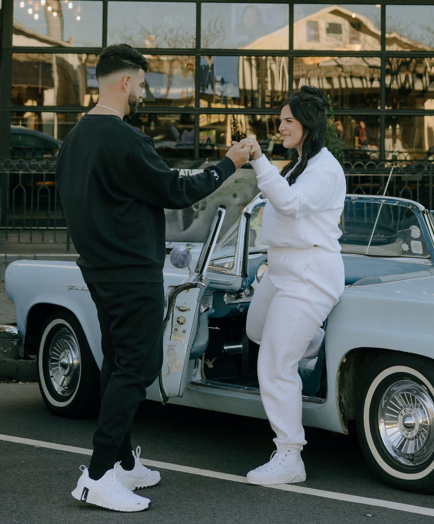 Two people in casual outfits interact playfully in a parking lot next to a classic car.