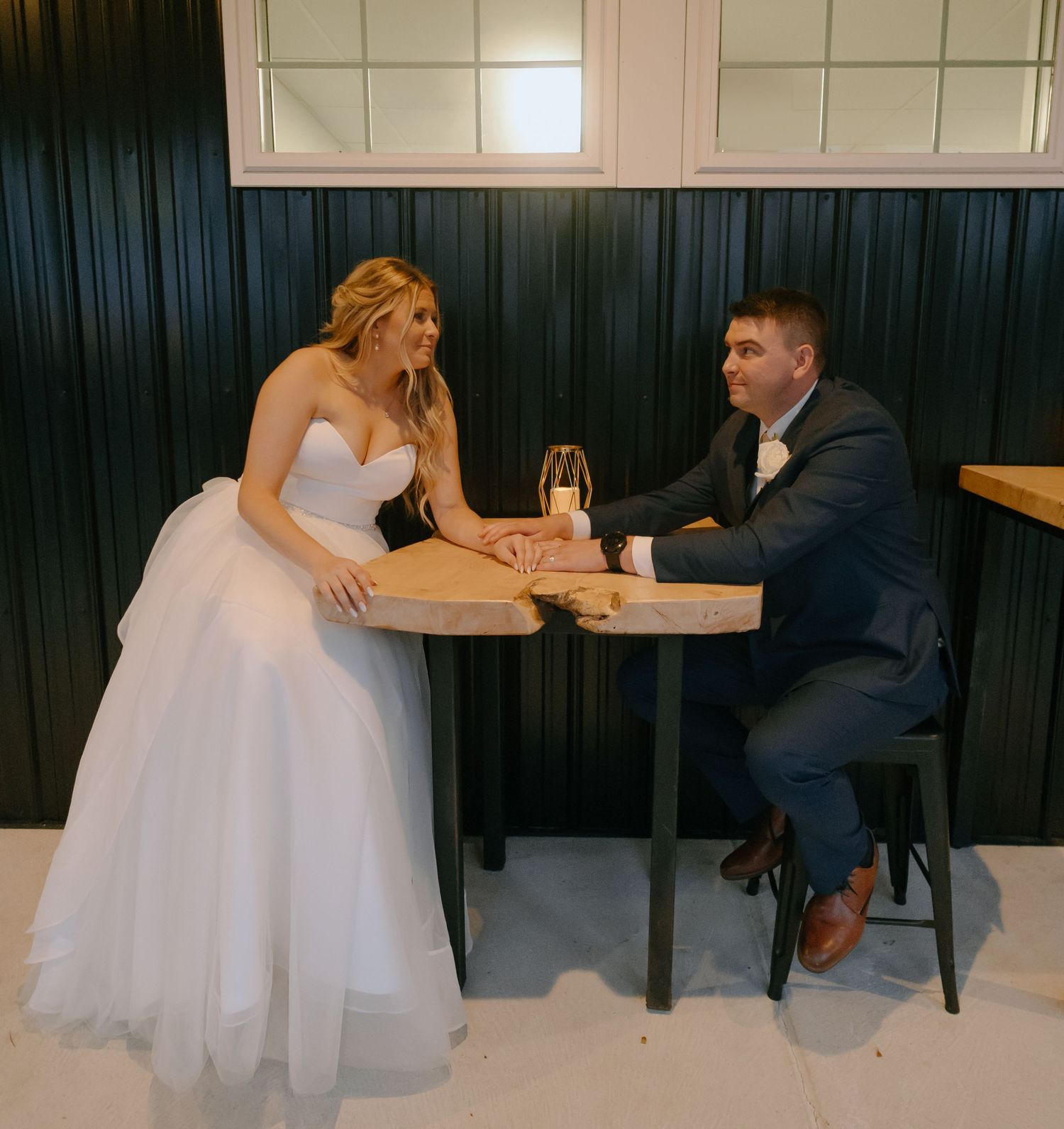 A bride in a white gown and groom in a suit share a casual moment at a small table with drinks.