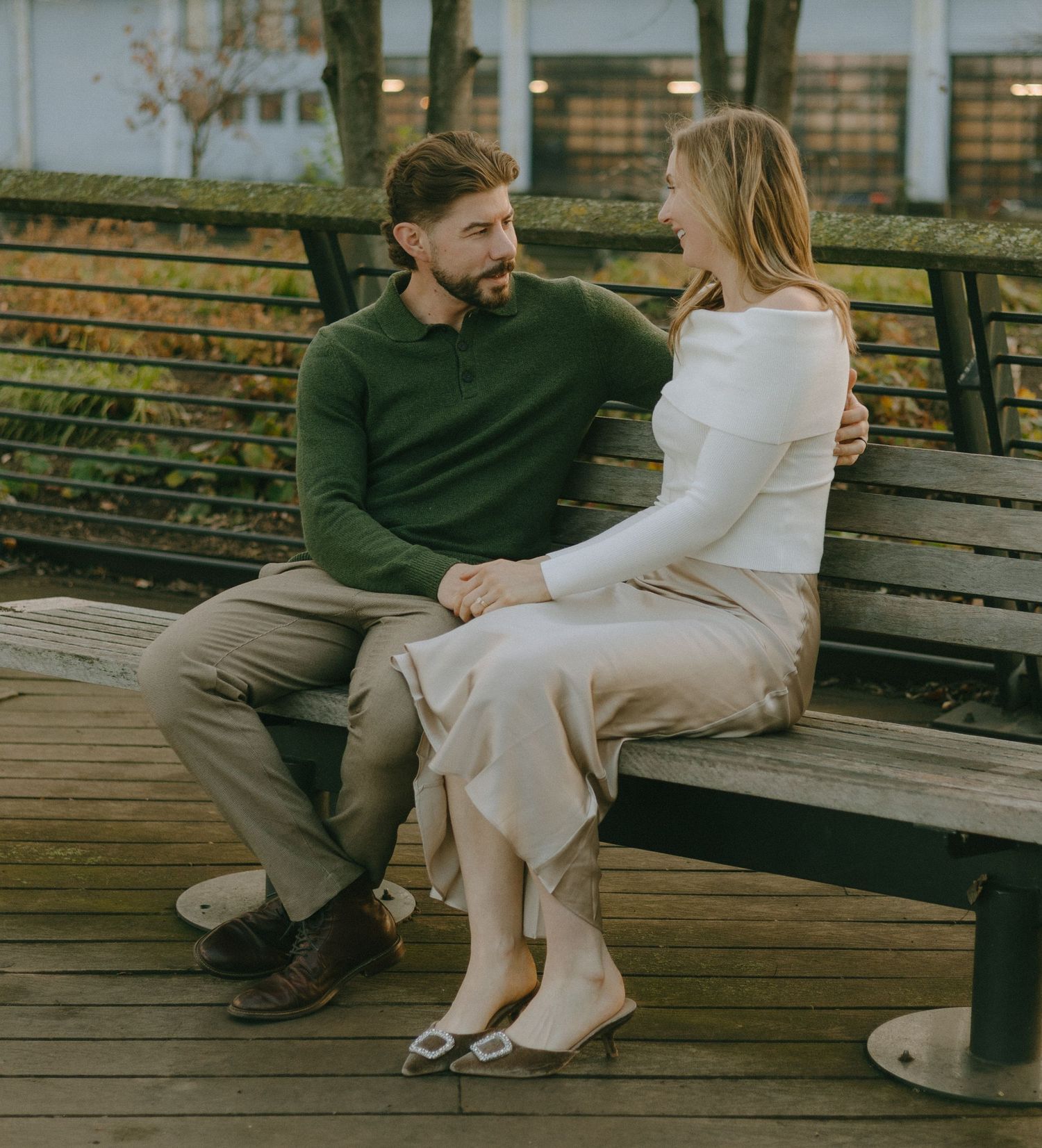 A couple in casual attire shares an intimate conversation while sitting together on a wooden bench outdoors.