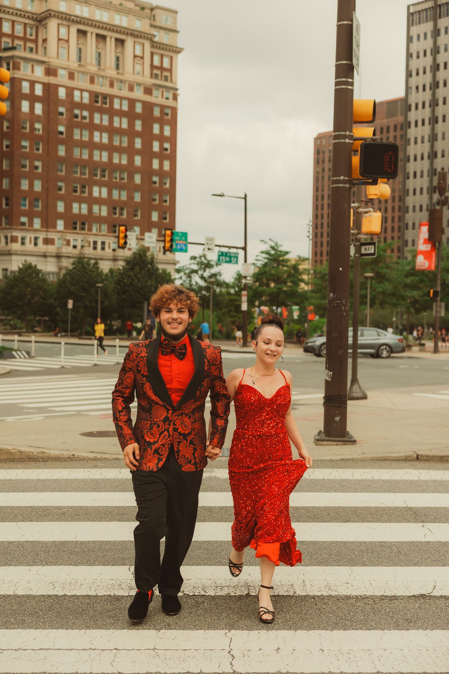A couple in coordinating red outfits walk across an urban street with brick buildings in the background.