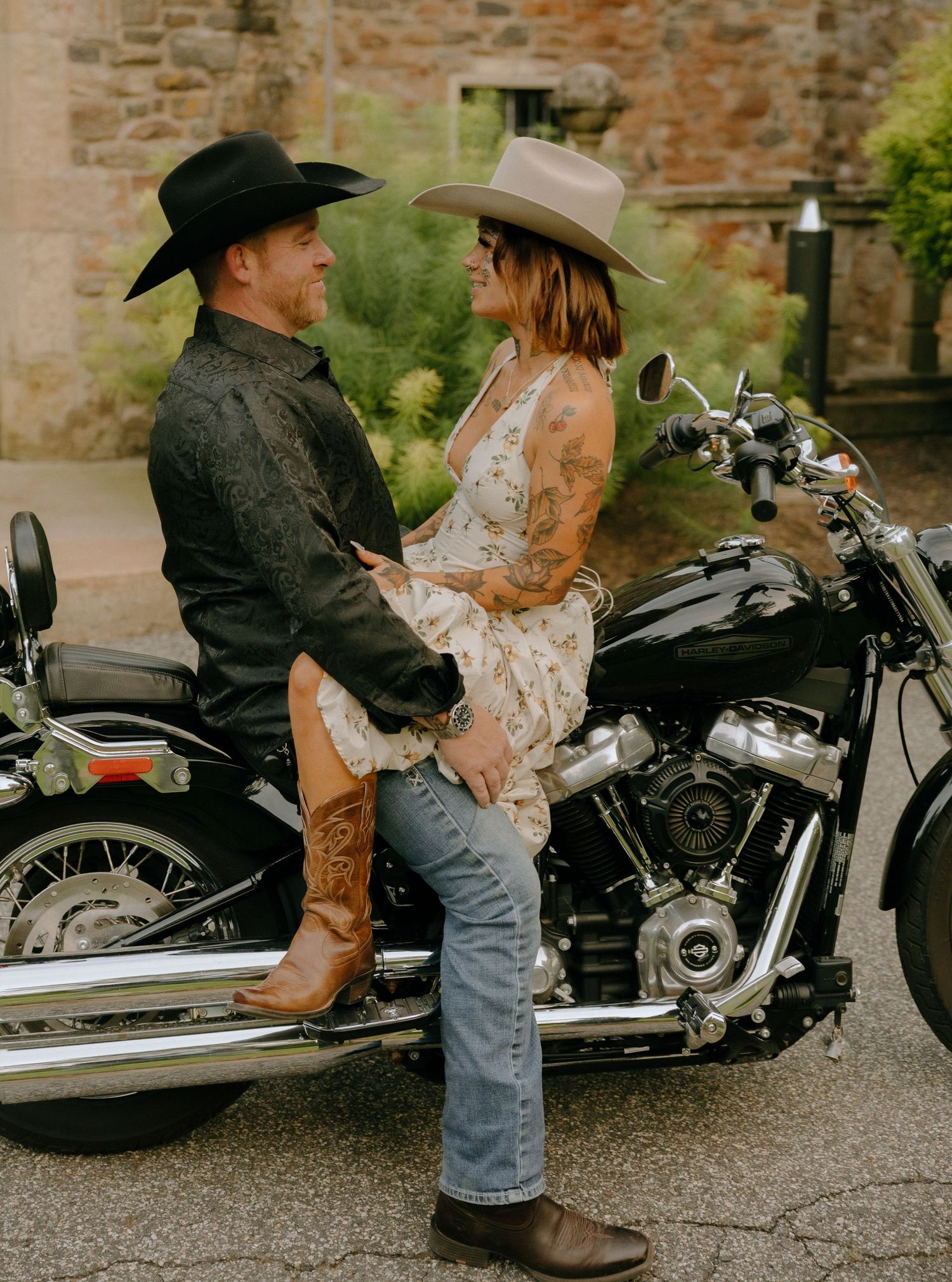Couple in western attire poses near a motorcycle against a rustic brick wall.