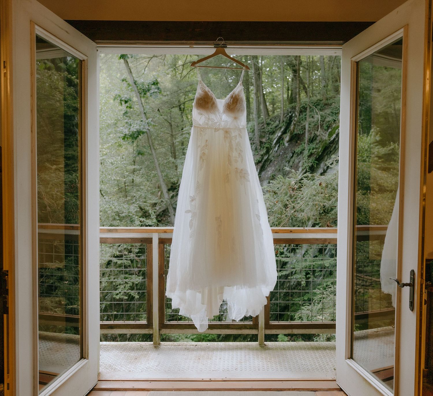 A white wedding dress hangs elegantly in a sunlit window-lined room with forest views.