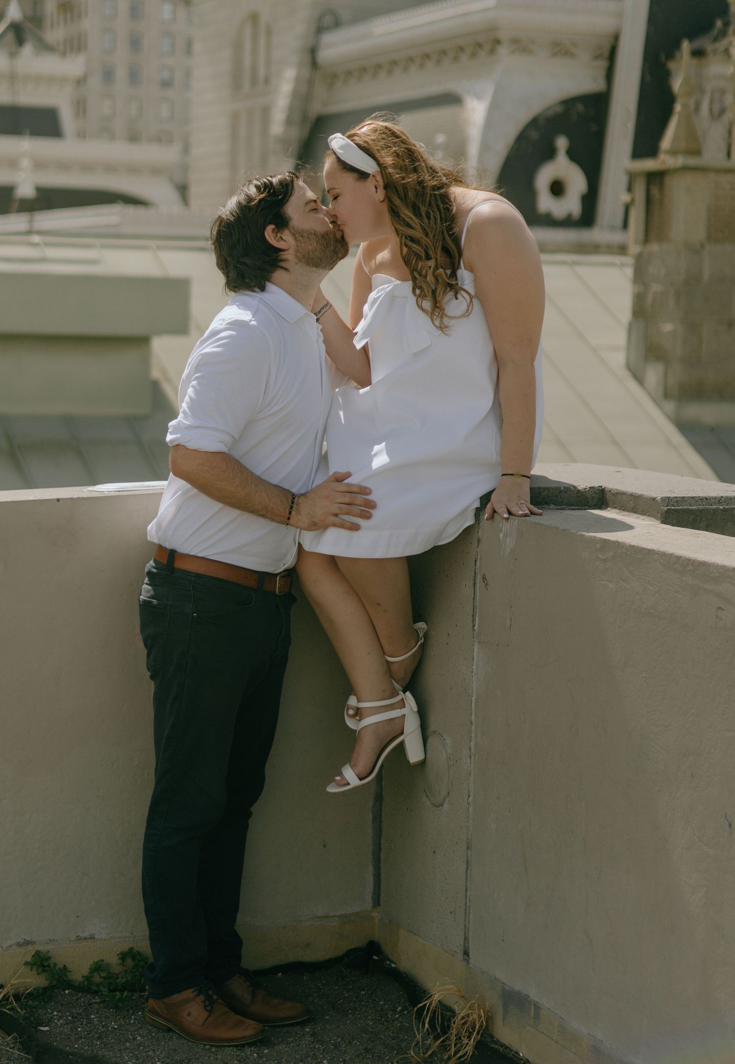 Couple embraces on concrete ledge with gothic architecture and city buildings in background.