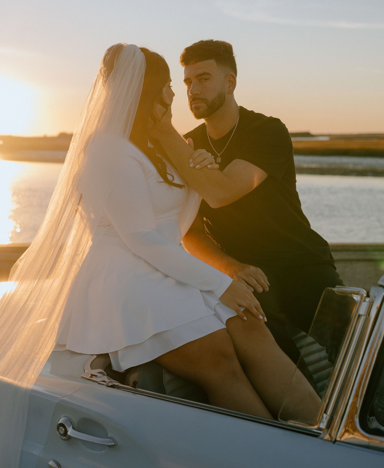 Romantic couple sitting on beach at sunset with flowing white fabric creating dreamy silhouettes.