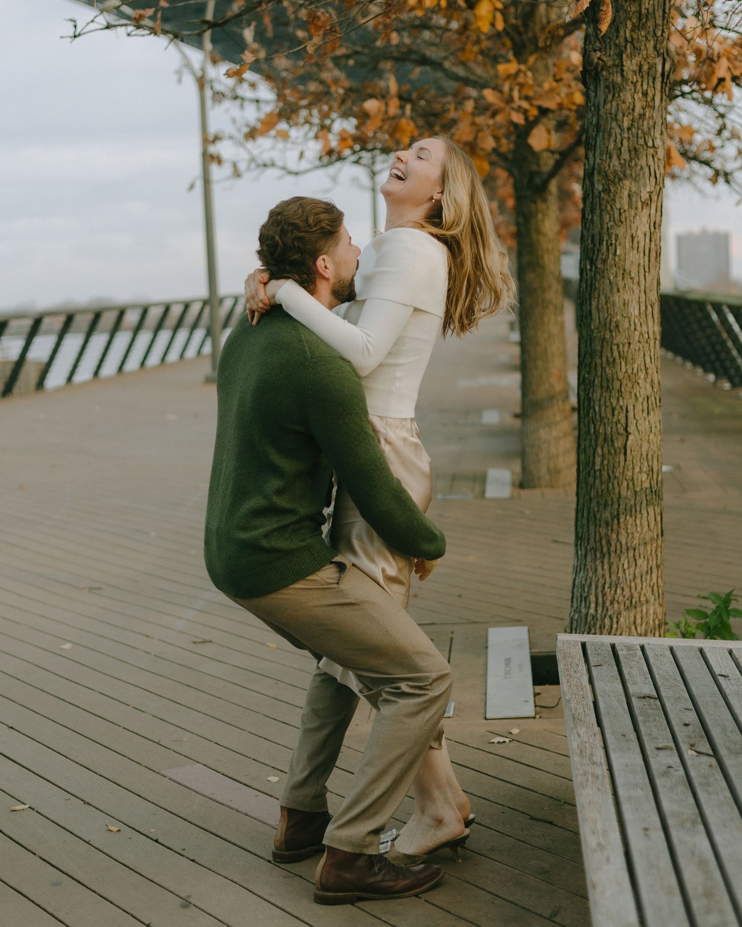 Two people share a romantic moment on a wooden boardwalk surrounded by autumn trees on a cloudy day.