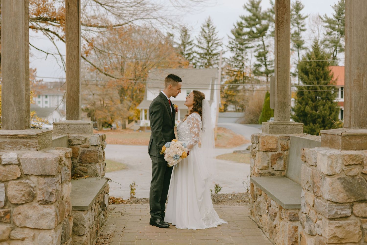Romantic winter wedding scene with couple embracing outdoors by rustic stone wall and bare trees.