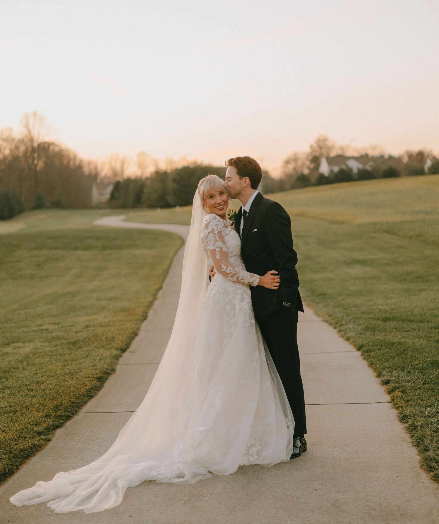 A couple shares an intimate moment at sunset on a concrete path surrounded by green lawns.