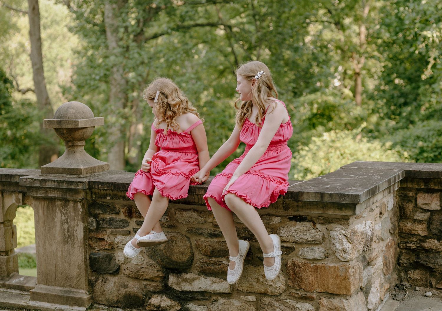 Two figures in bright pink dresses sit together on a stone wall in a garden setting.