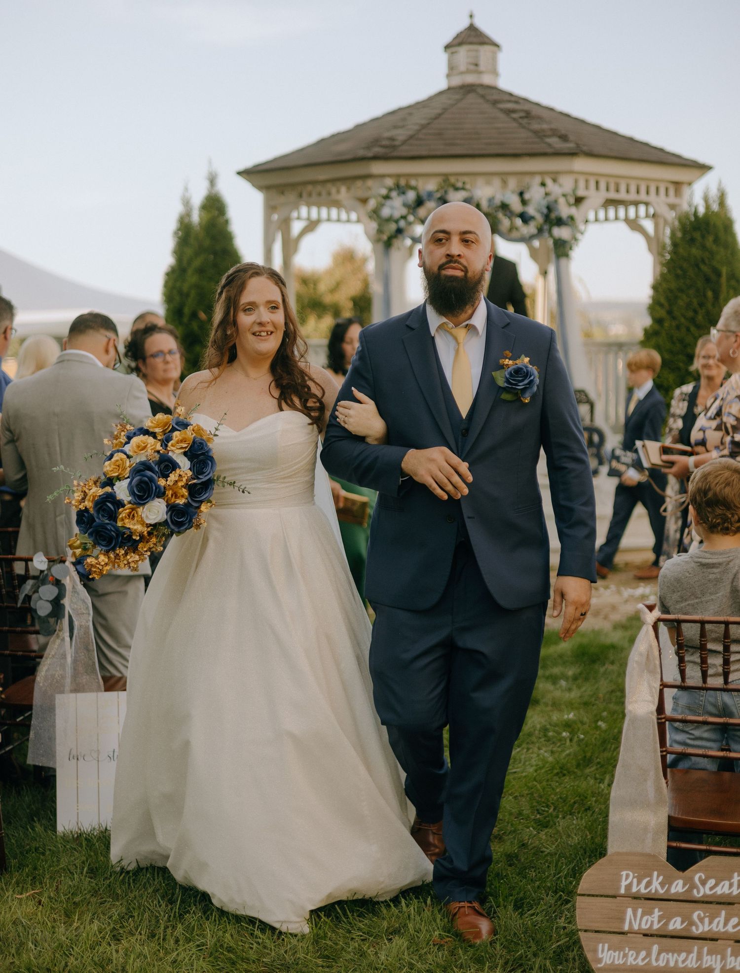 A wedding ceremony setup with a gazebo, wooden signs, and sunflower bouquets on a grassy lawn.
