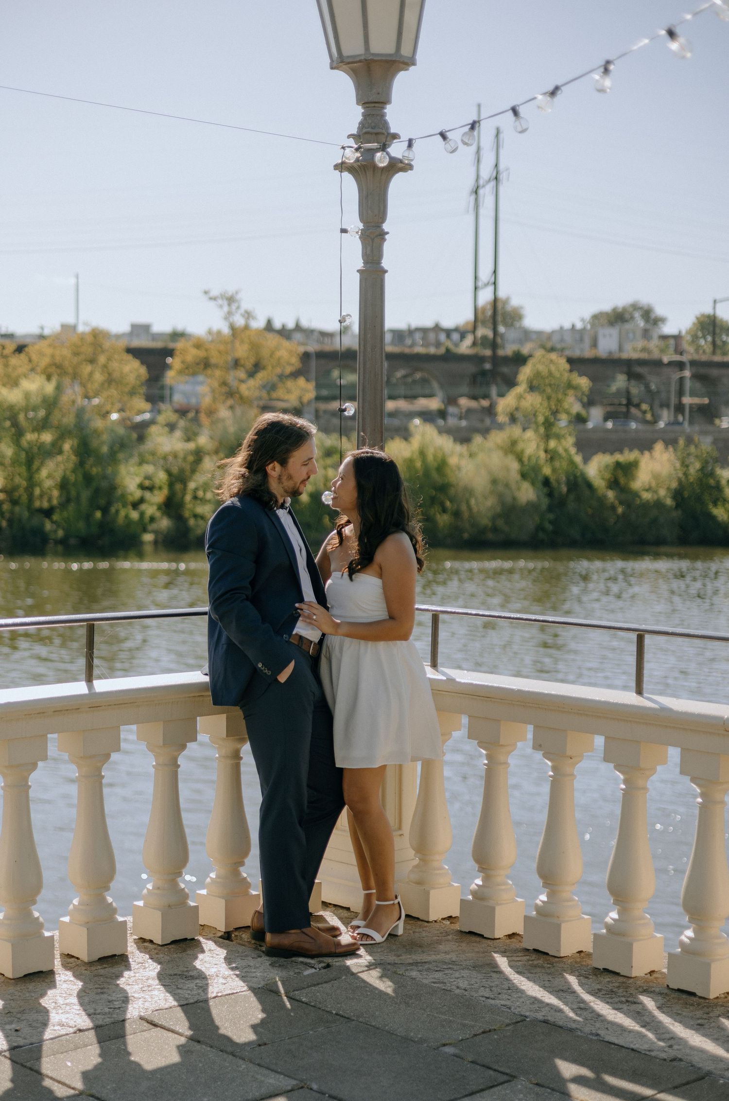 A romantic couple shares a tender moment on an ornate balcony overlooking a lake with autumn trees in the background.