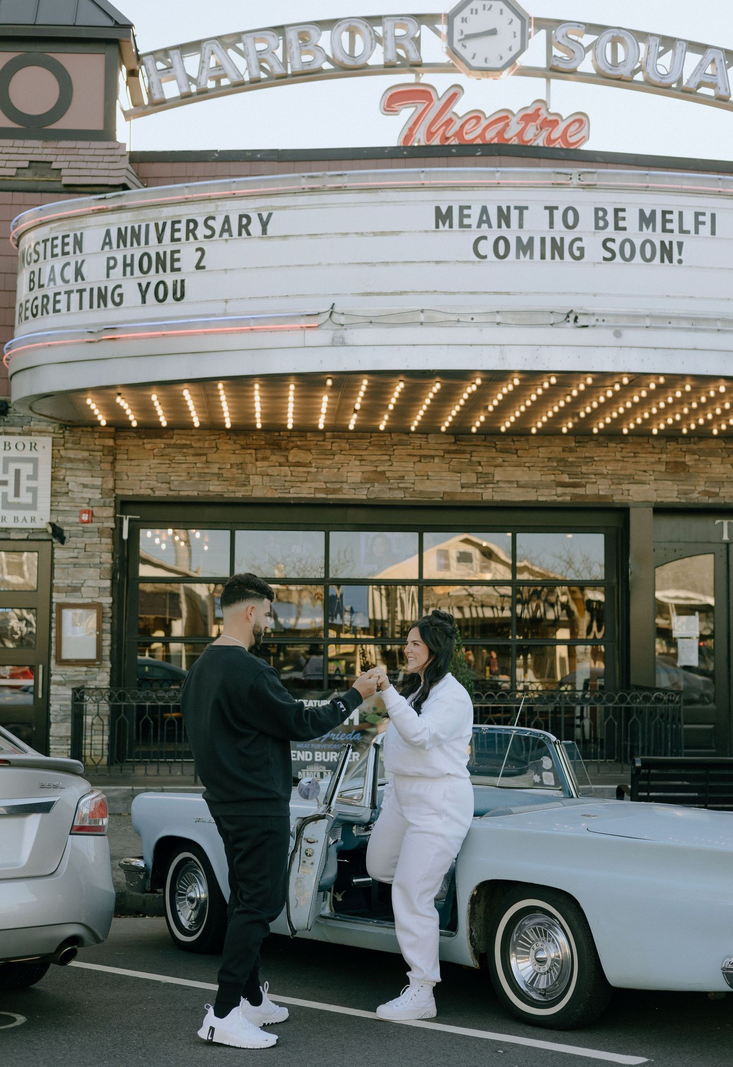 Vintage movie theater exterior with illuminated marquee and classic white car parked outside.