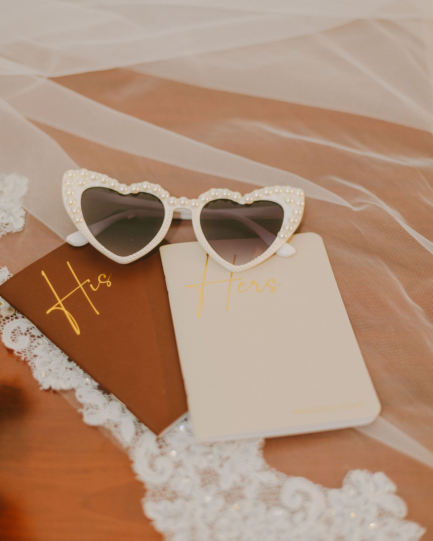 White heart-shaped sunglasses and wedding vow cards rest on a wooden surface with delicate lace veil.