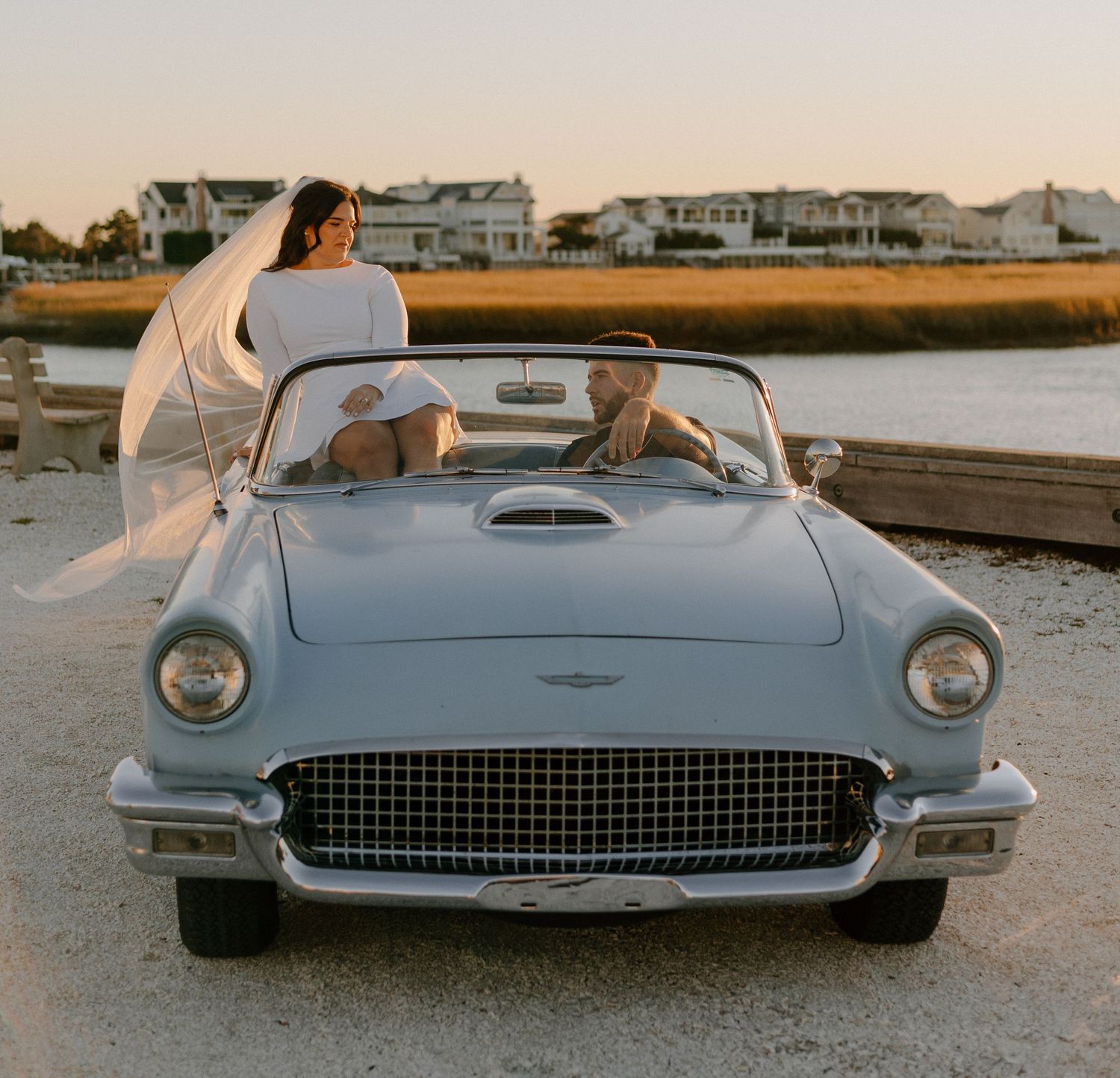 A classic light blue Ford Thunderbird convertible parked by the water at sunset with occupants enjoying the coastal view.