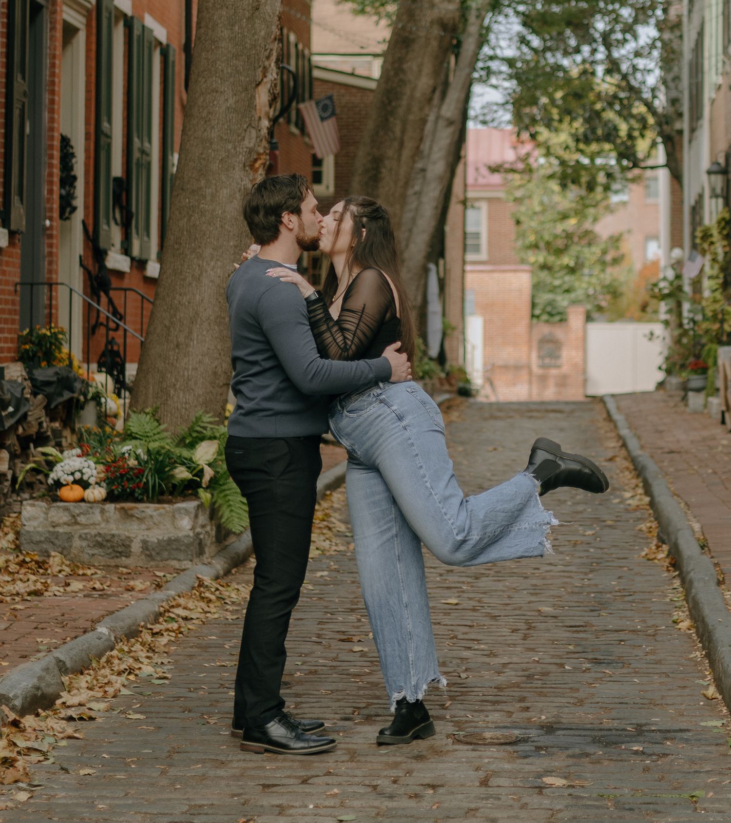 A couple shares a romantic moment on a cobblestone street lined with autumn leaves and historic buildings.