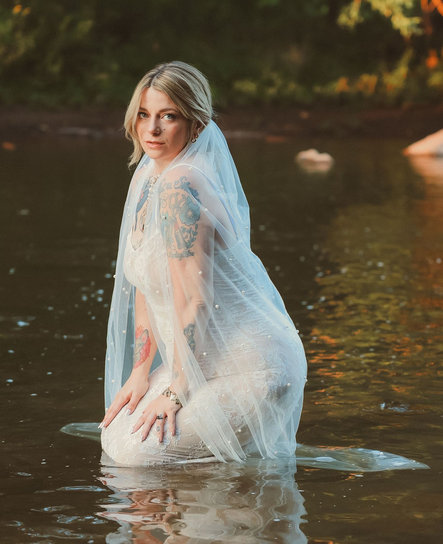 A bride poses dramatically in shallow river water while showing off her wedding dress and veil.