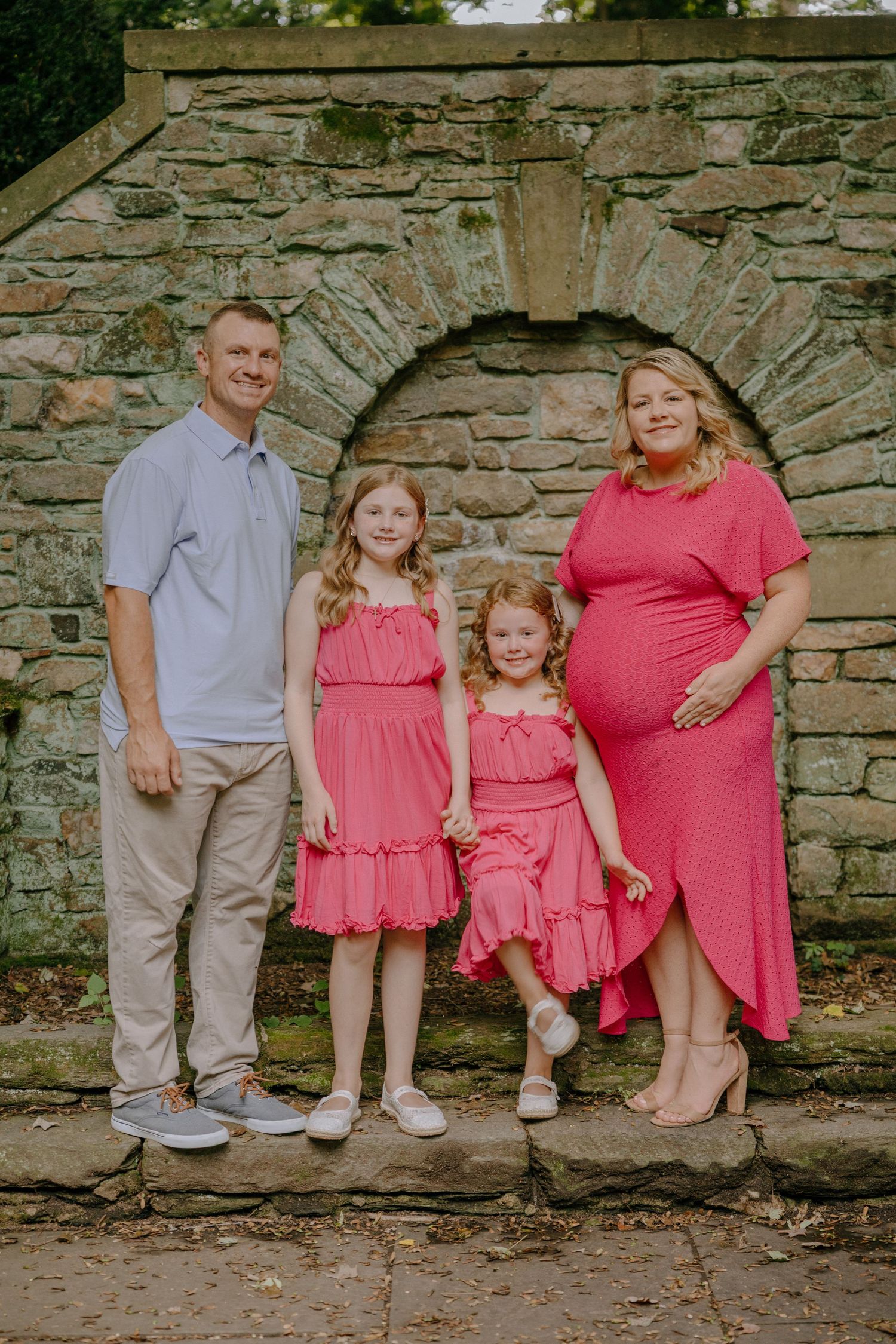 A family poses along a rustic stone wall wearing coordinated pink and gray outfits for a casual outdoor photoshoot.
