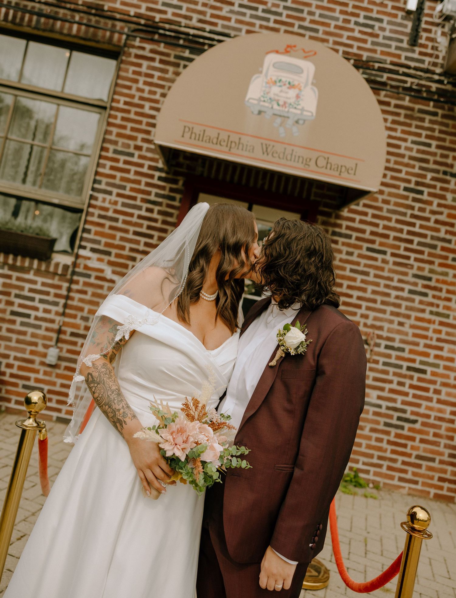 Wedding ceremony outside a brick chapel with floral arrangements and a red velvet rope.