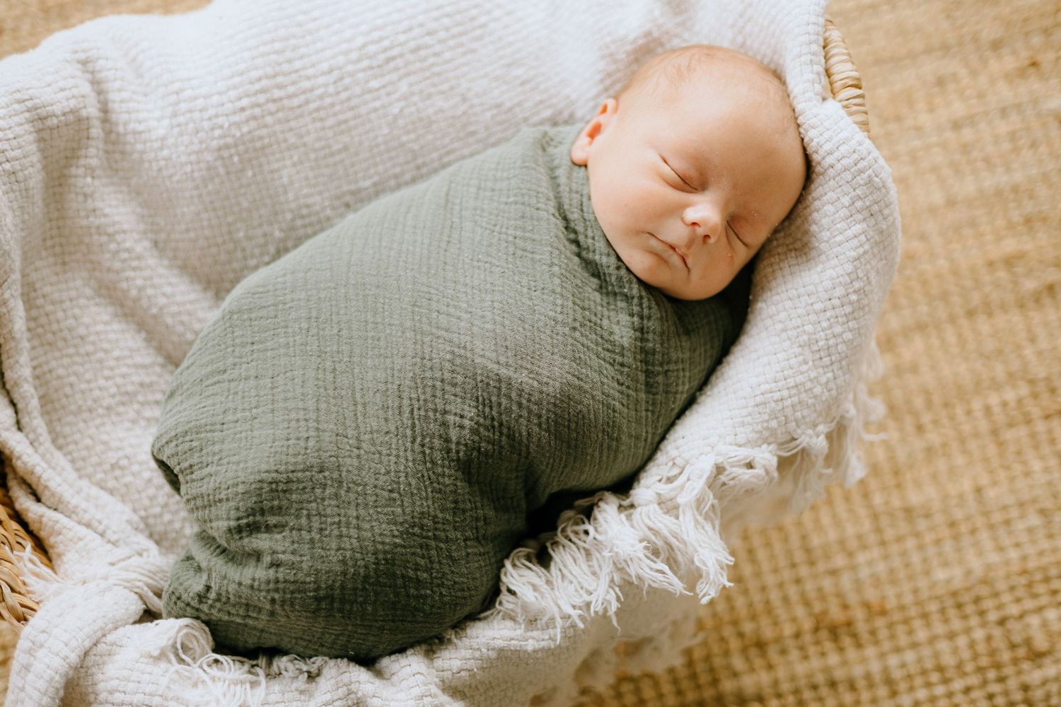 A sleeping newborn baby swaddled in white fabric lies on a textured beige blanket.