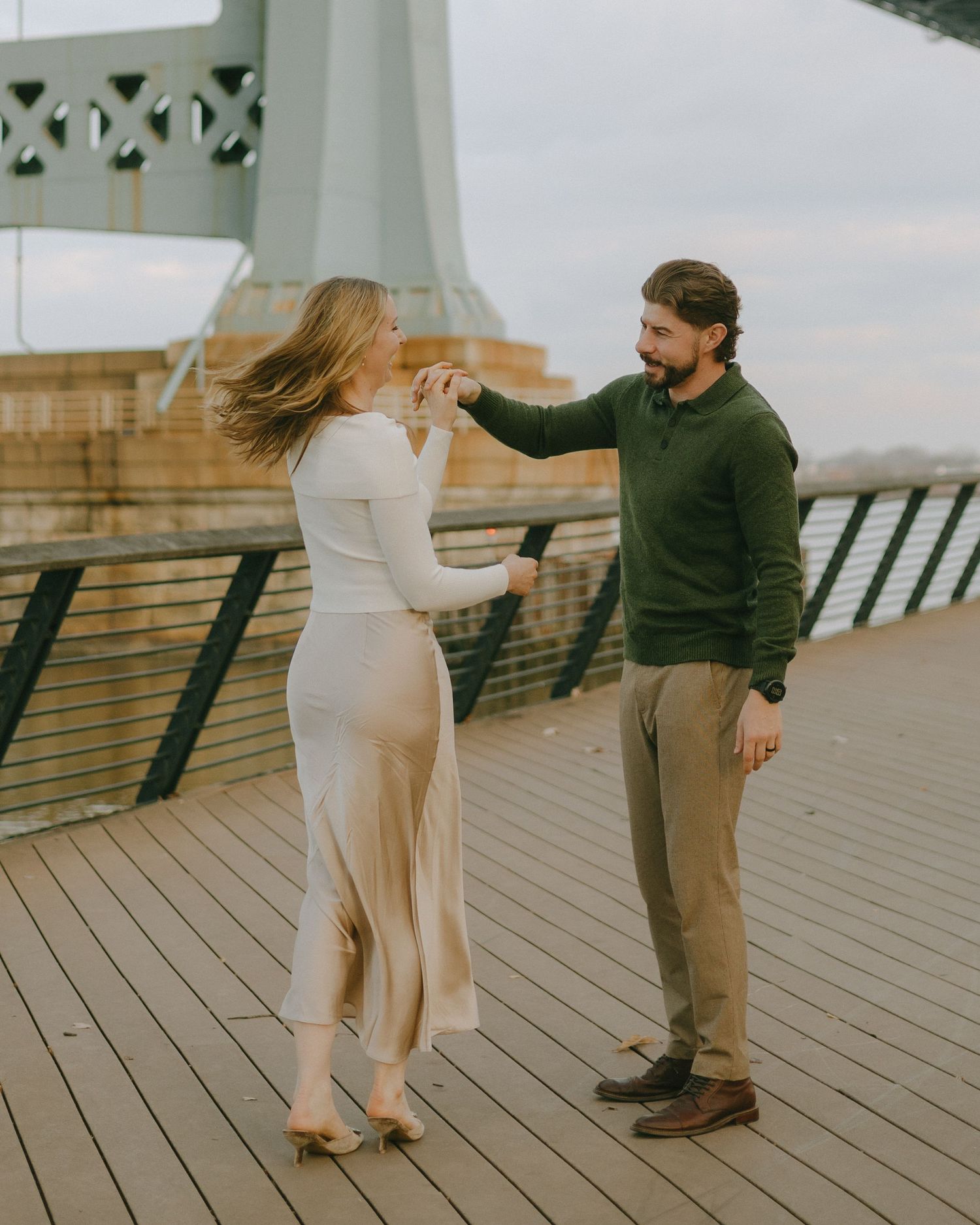 A playful couple dancing and laughing together on a waterfront boardwalk under a bridge.